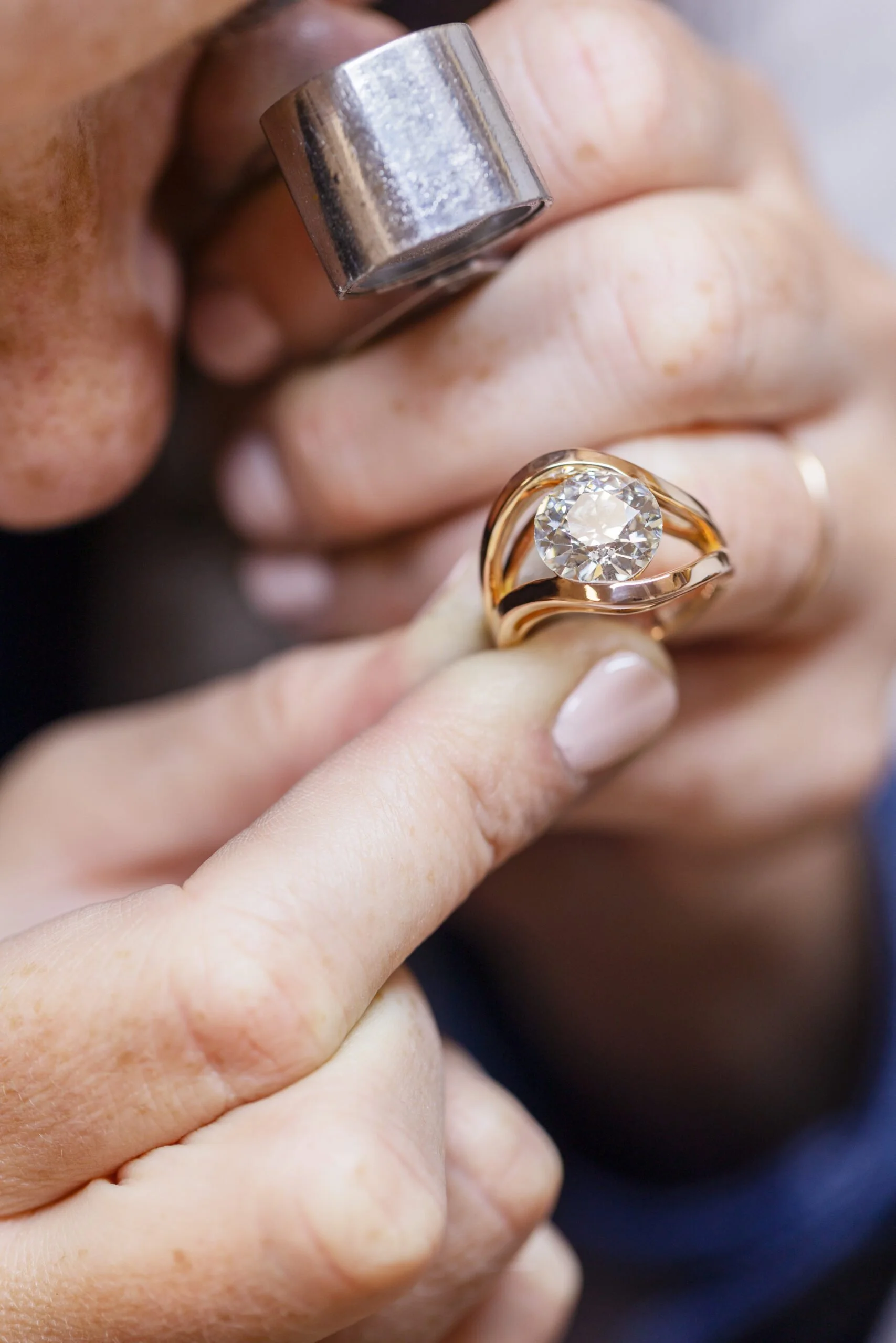 Gros plan d'une main portant une bague en or avec un grand diamant au centre, tenant un objet métallique cylindrique près d'une autre main avec des imperfections cutanées