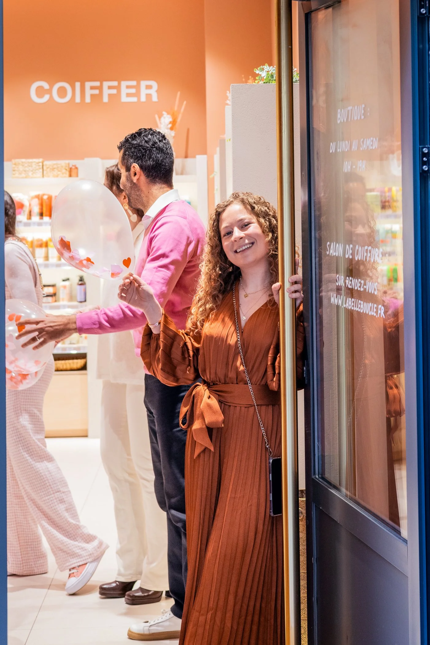 Une jeune femme souriante de cheveux bouclés qui tient une bulle de savon dans une main, se tient à la porte d'un salon de coiffure. À l'intérieur, plusieurs personnes discutent, avec un mur orange en arrière-plan portant le mot 'COIFFEUR'.