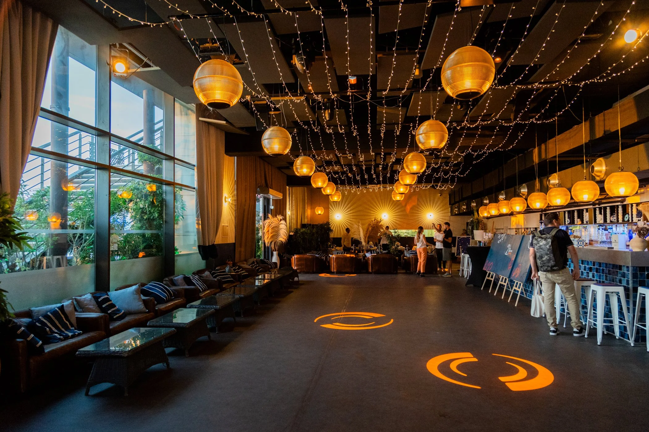Intérieur d'un café ou restaurant avec des lampes suspendues dorées et des guirlandes lumineuses, des fenêtres larges laissant entrer la lumière naturelle, et des personnes en train de discuter au comptoir.