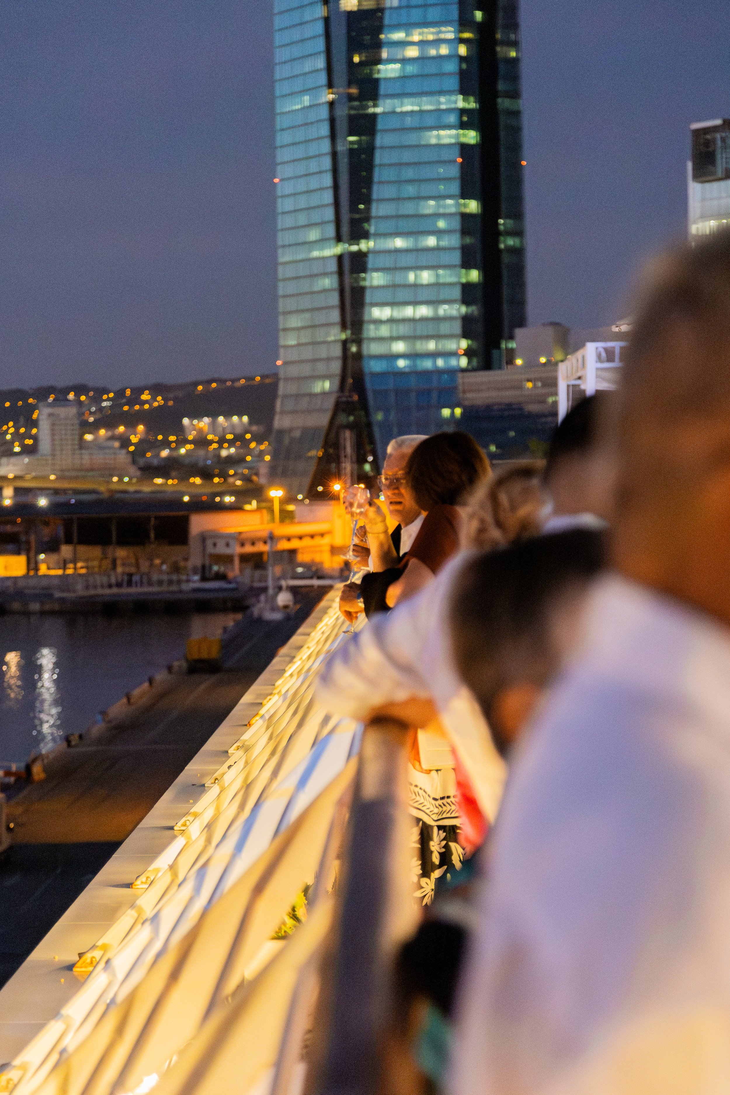 Groupe de personnes regardant un paysage urbain la nuit, avec un gratte-ciel en verre ilumines et des lumières de ville en arrière-plan.