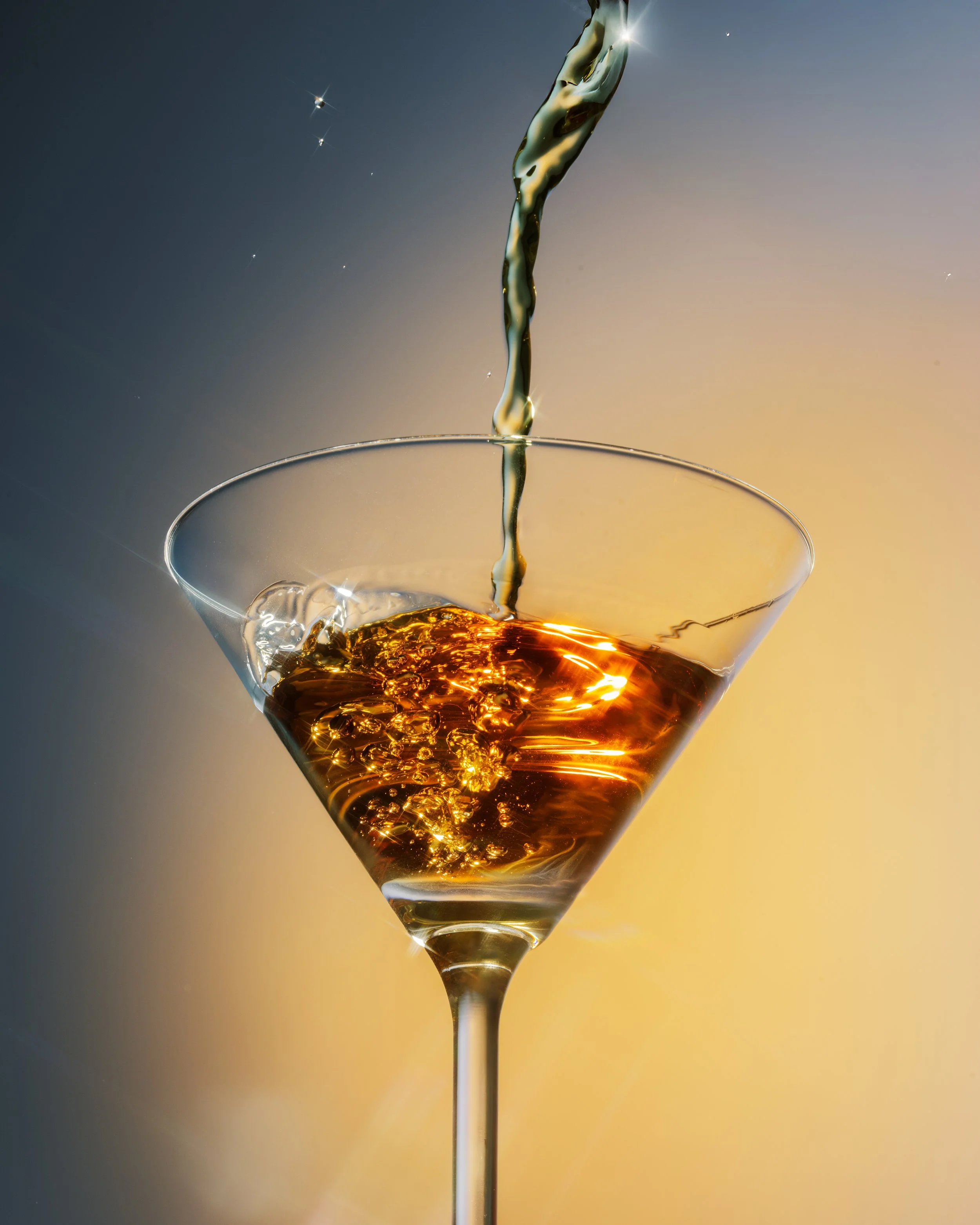Un liquide ambré est versé dans un verre à cocktail, avec un fond dégradé de couleurs chaudes et froides.