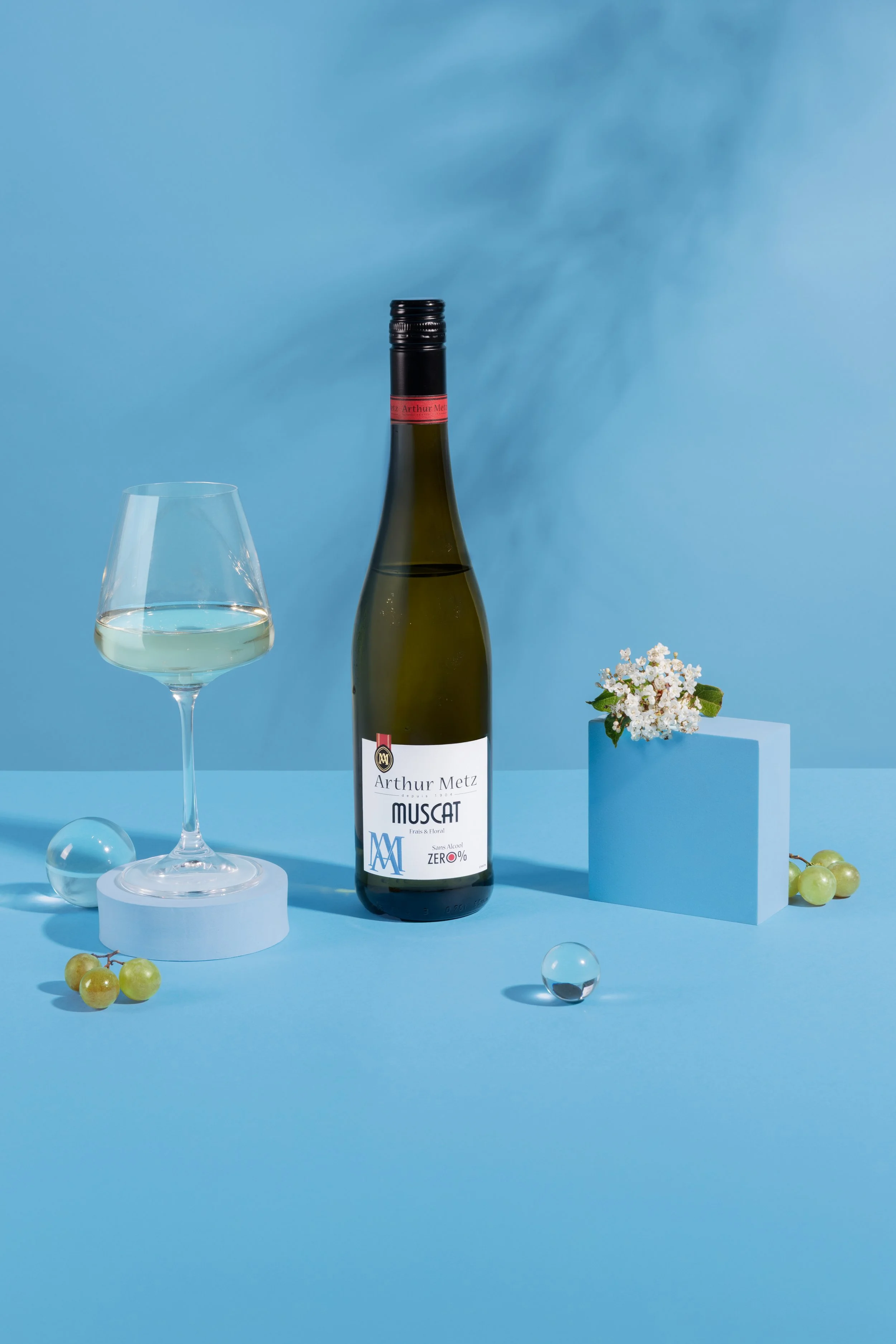 Une bouteille de vin blanc Arthur Metz Muscat avec un verre à vin blanc rempli à côté, un petit bouquet de fleurs blanches, des grappes de raisin vert, quelques boules en verre translucides, le tout posé sur une surface bleue avec un fond bleu.