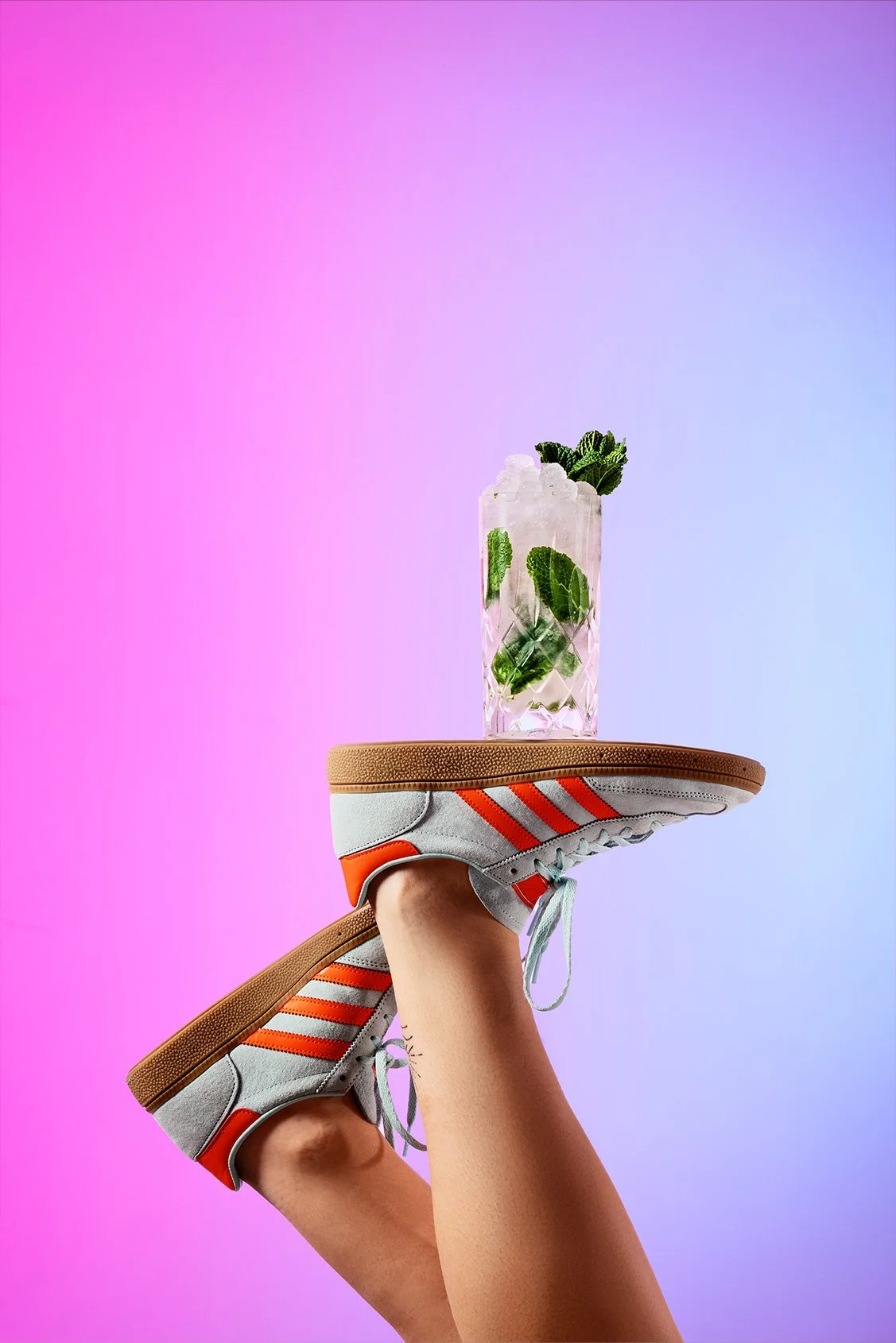 Un pied chaussé de baskets blanches avec des rayures orange, tenant un fauteuil avec une chaussure sur la jambe. La chaussure supporte un verre de cocktail contenant de la glace et de la menthe, contre un fond dégradé de rose et violet.