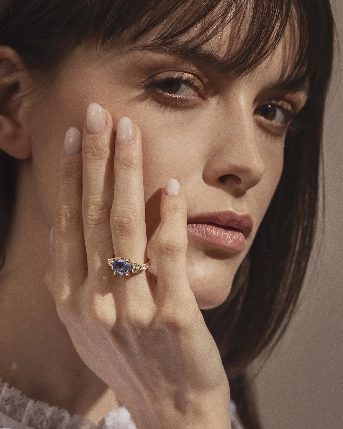 Une femme regarde l'objectif, portant une bague avec un grand saphir bleu entouré de diamants et un maquillage naturel, tenant sa main près de son visage.