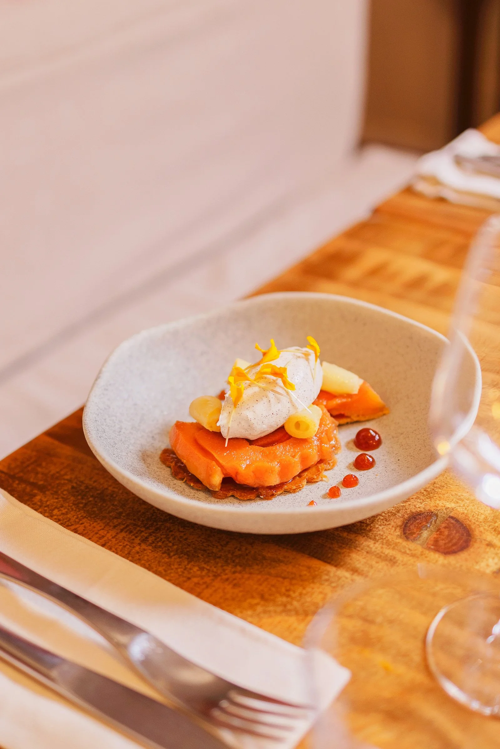 Une assiette contenant un plat avec du saumon, une sauce crémeuse, une quenelle de crème, décorée de zestes d'orange et de petites gouttes de sauce orange, sur une table en bois.