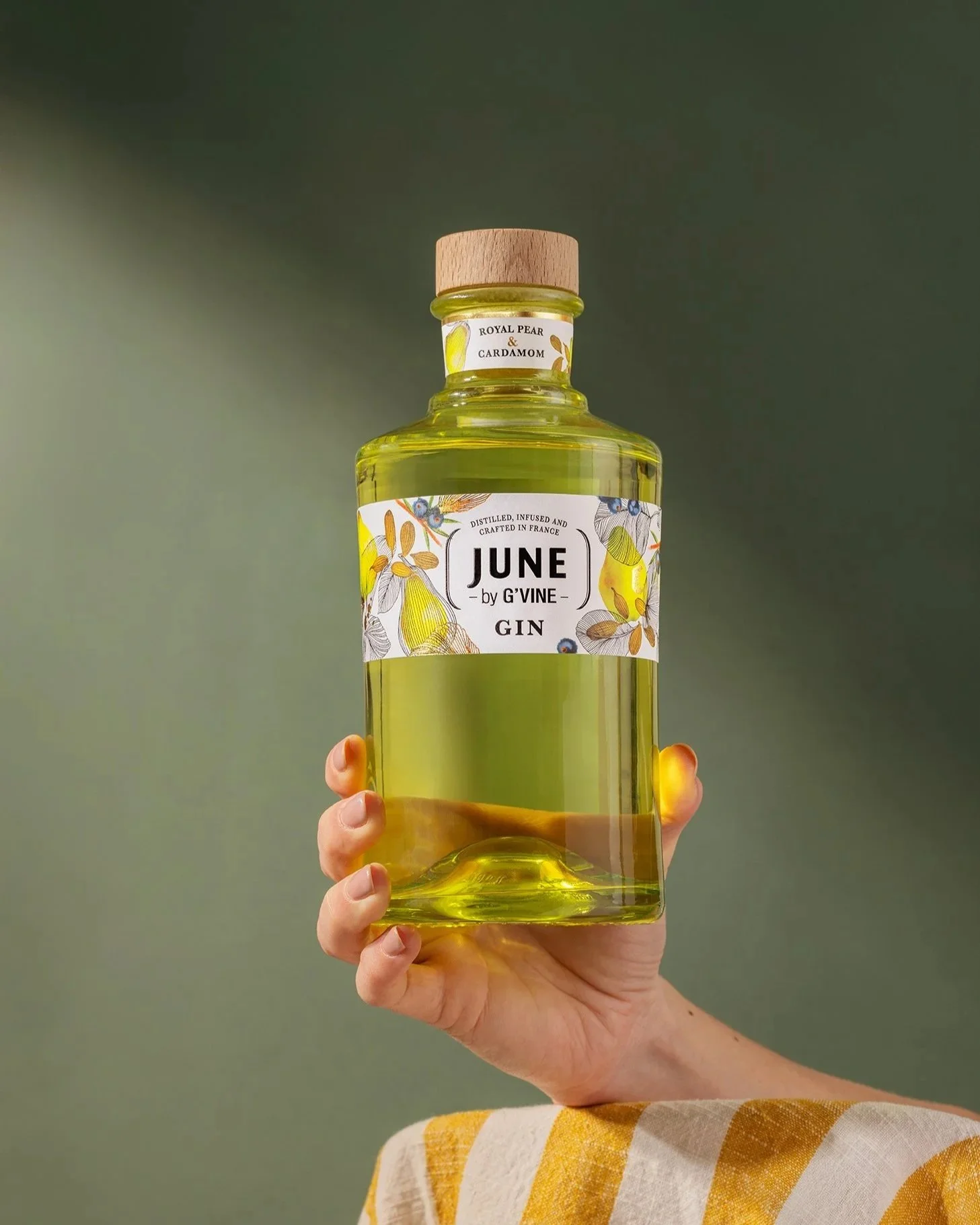 Bouteille de gin infusé avec des cerises royales et du cardamome, tenue par une main, avec un fond vert.