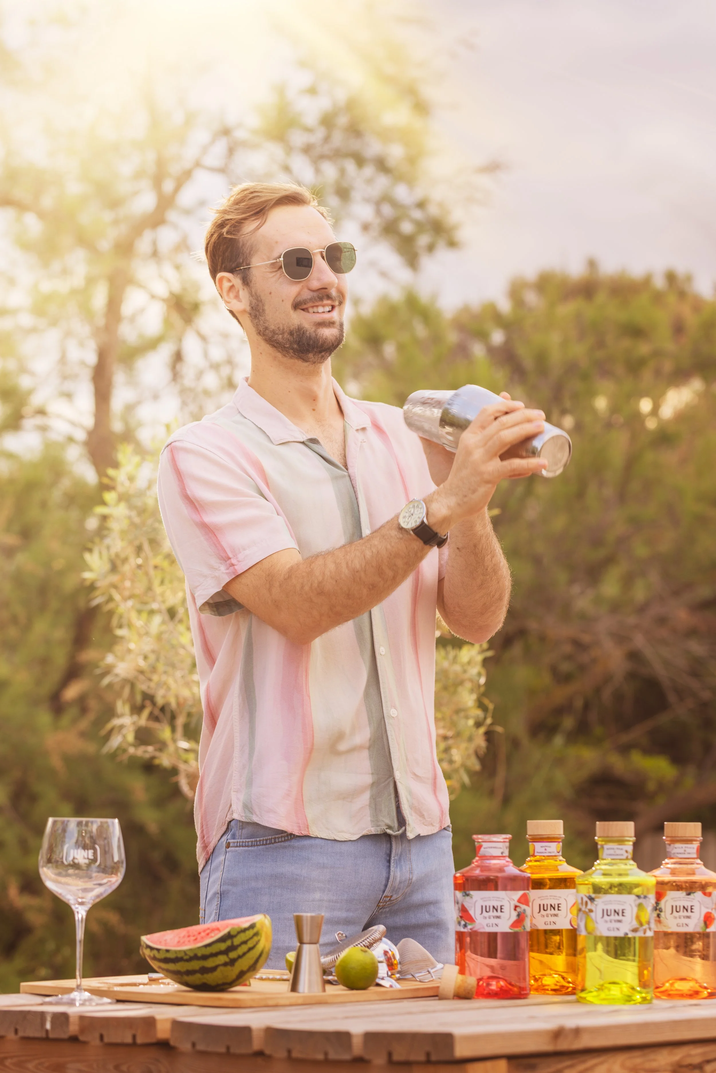Un homme avec des lunettes de soleil, une barbe, portant une chemise à rayures pastel, se tient devant une table en plein air avec plusieurs bouteilles de gin colorées, un verre à vin, une tranche de pastèque, un melon, des pommes vertes, et des ustensiles de cuisine.
