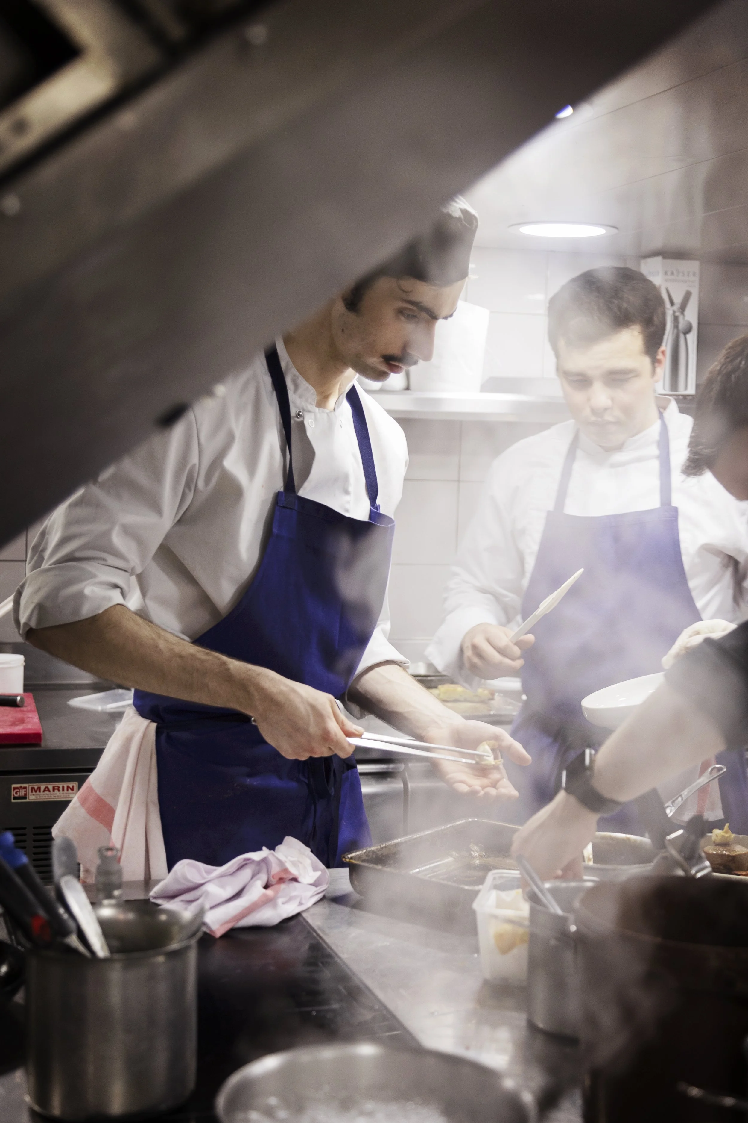 Deux chefs en uniforme cuisinent dans une cuisine professionnelle. L'un d'eux est en train de préparer un plat, l'autre regarde attentivement. Il y a des ustensiles et des casseroles autour d'eux.