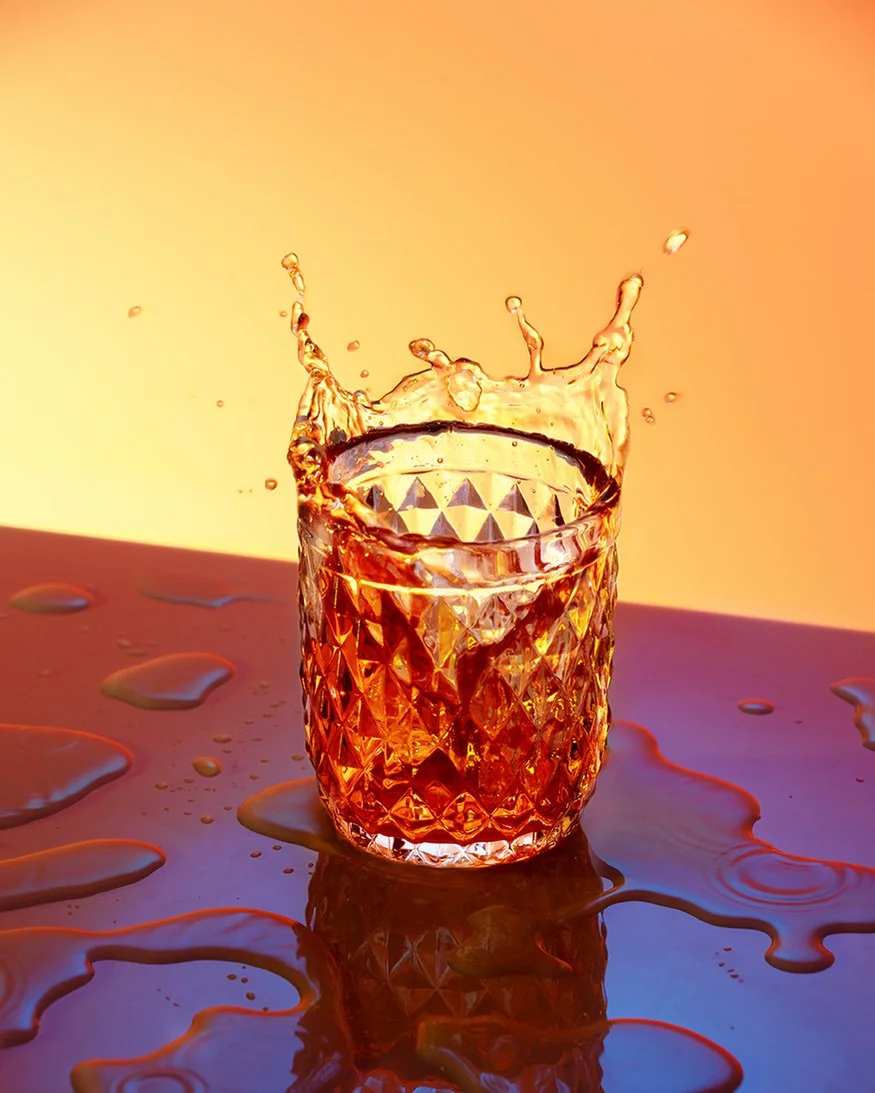 Verre en verre avec une boisson ambrée, éclaboussures d'eau autour, fond chaud.