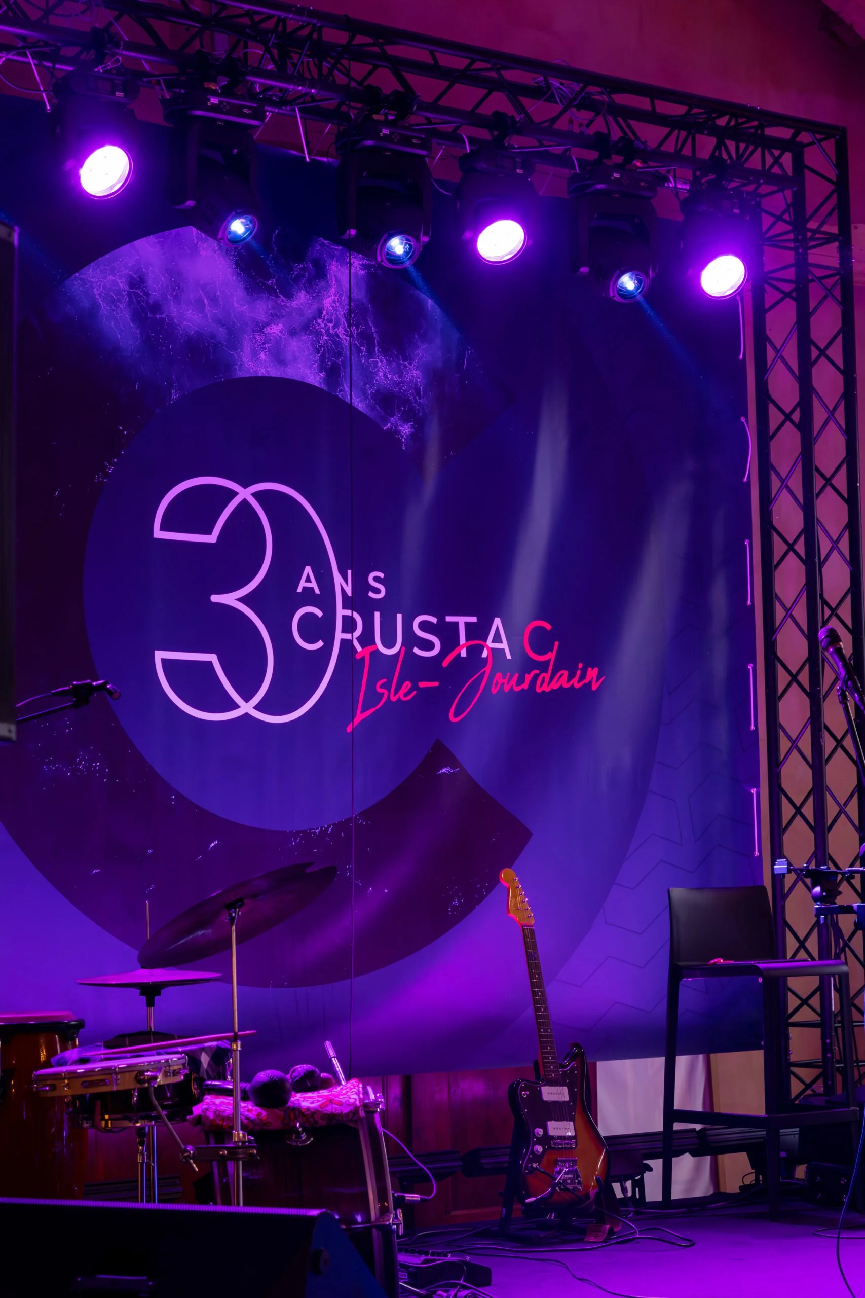 Scène de scène de concert ou d'événement avec éclairage violet, guitare électrique, batterie, un fauteuil, et un mur avec un logo indiquant 30 ans d'Acoustique Île-Jourdain.