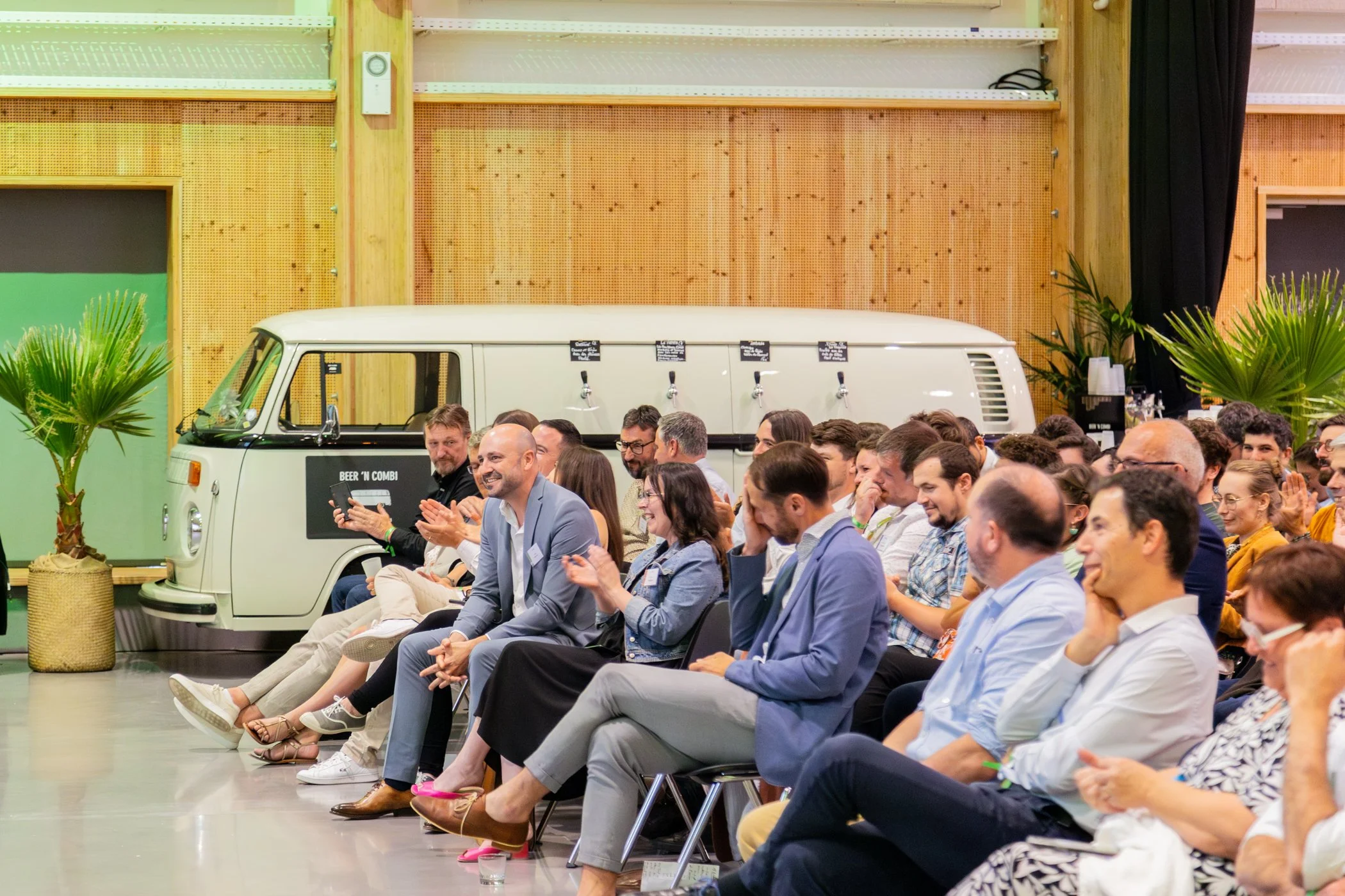 Une foule de personnes assises dans un auditorium, certains applaudissant, entourées de plantes vertes et d'un décor en bois avec un camion blanc en arrière-plan.