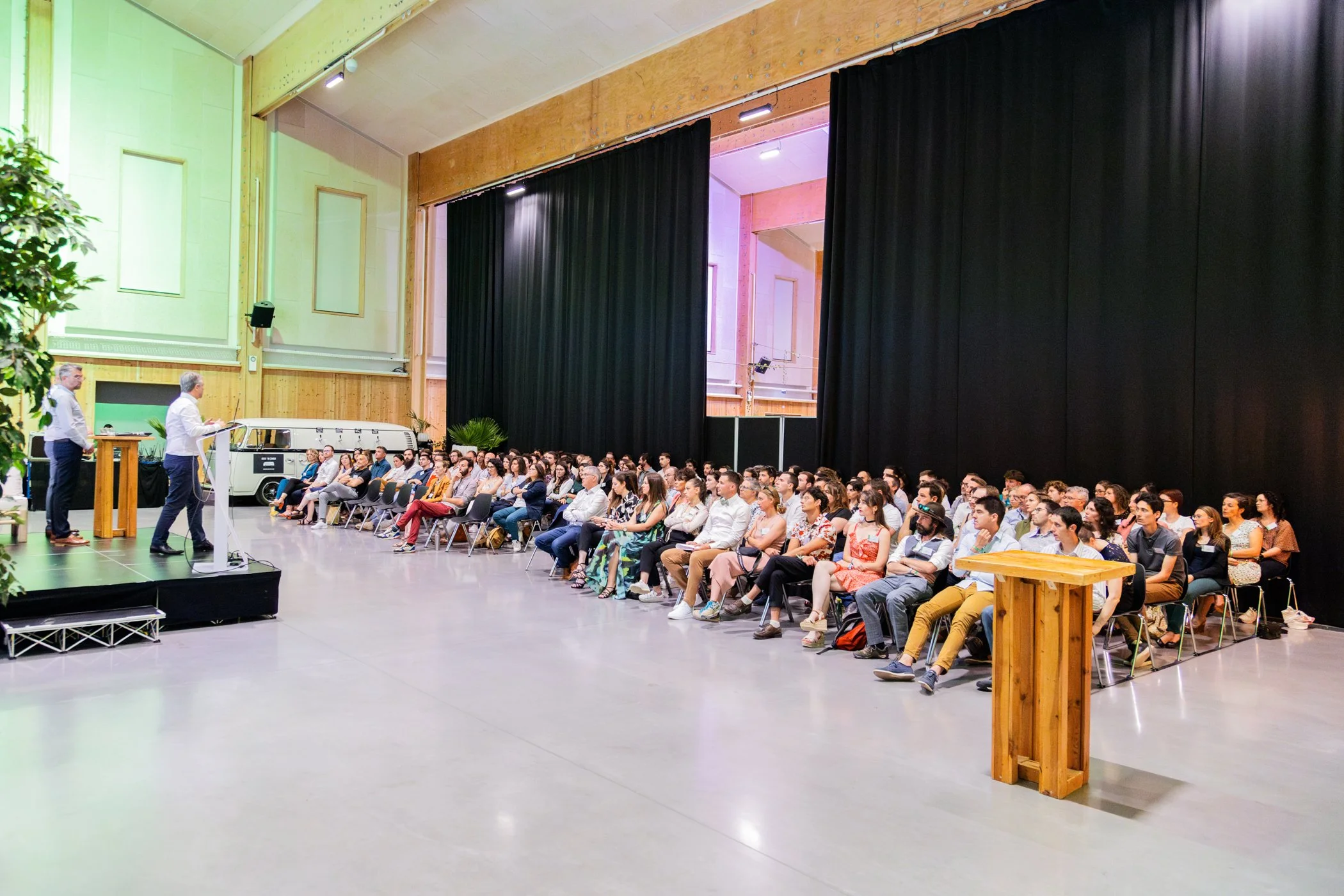 Une grande salle avec un public d'environ une centaine de personnes assises en rangées, assistant à une conférence ou présentation. Deux hommes en chemise blanche se tiennent à une estrade devant le public, avec un ordinateur portable. Il y a un gran