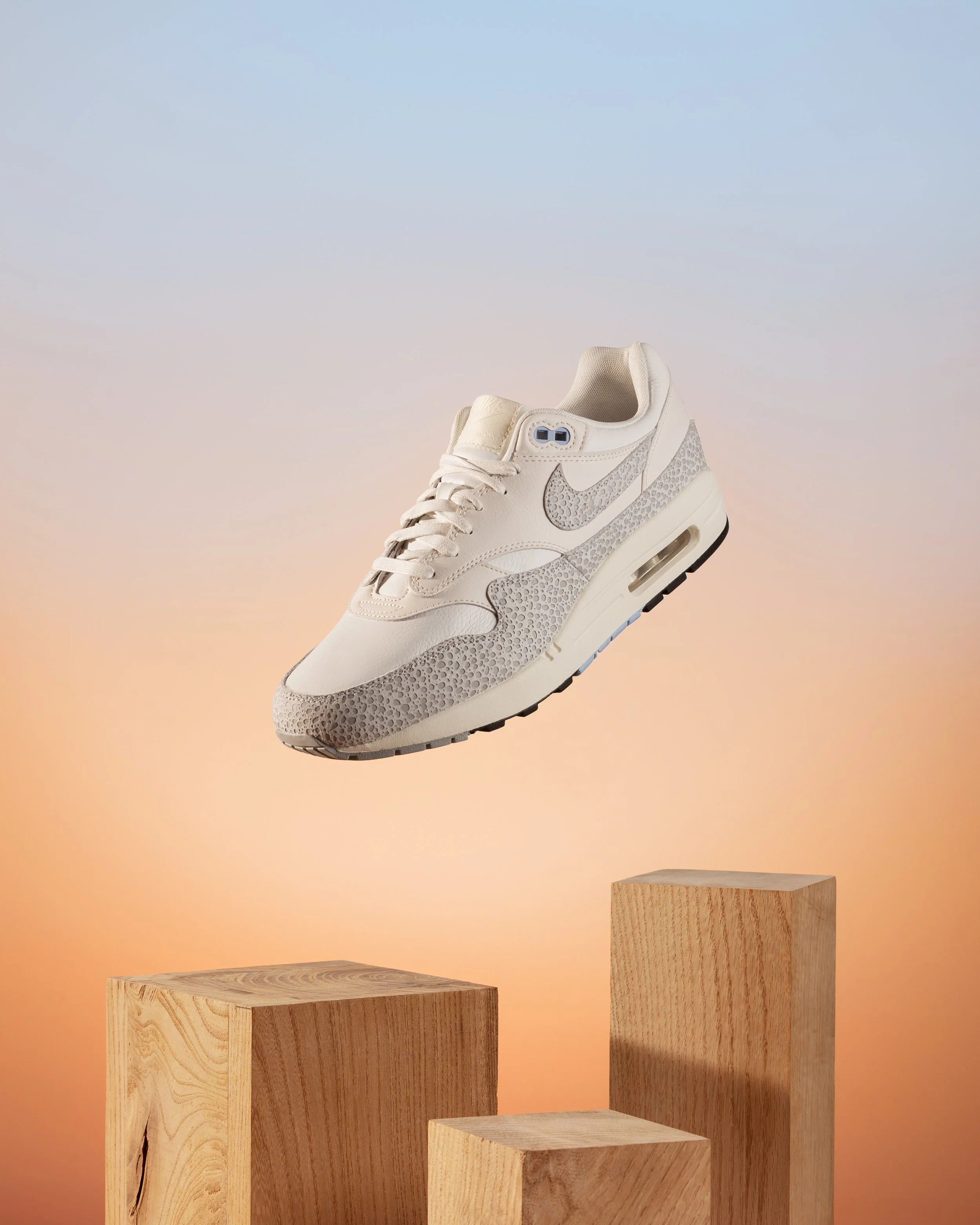 Une chaussure de sport blanche avec des détails en gris, posée sur une plateforme en bois, contre un fond dégradé de couleurs pastel du bleu au orange.
