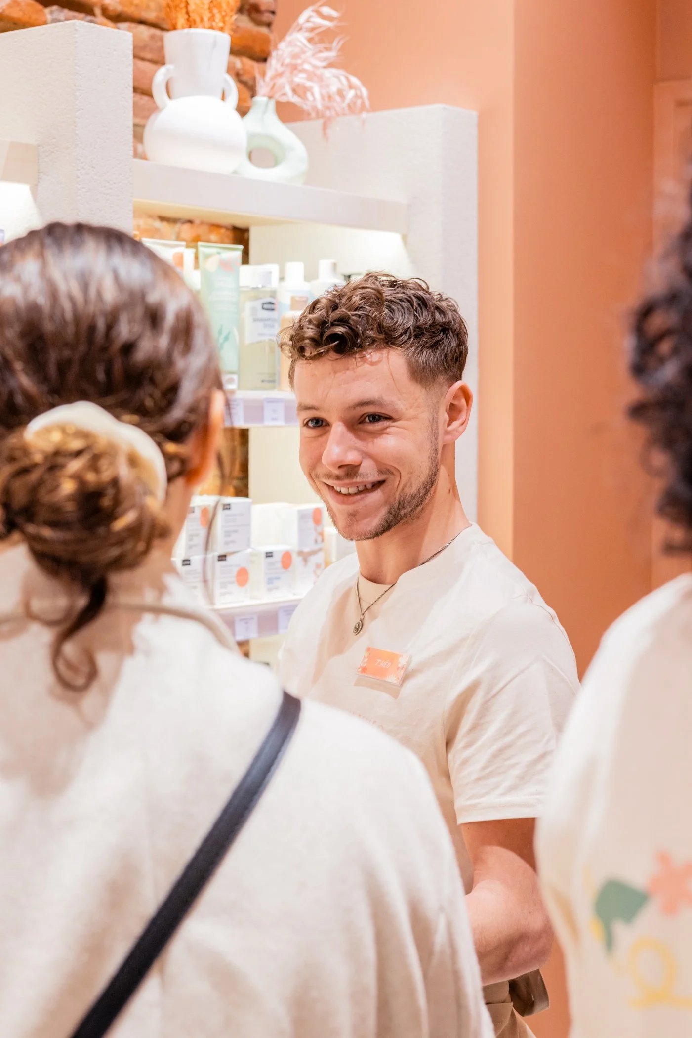 Jeune homme souriant porteur d'un t-shirt blanc, entouré de deux femmes dans un espace intérieur avec des étagères de produits de soins ou cosmétiques.