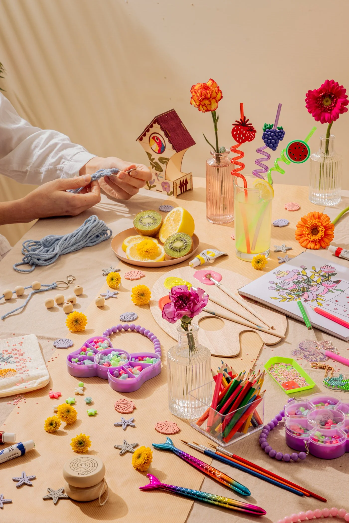 Table colorée avec des fleurs, fruits, perles, jouets, coloriages, matériaux pour création et décoration, comprenant des pinceaux, crayons, rubans, lots de perles, fleurs artificielles, pagelines, etc.