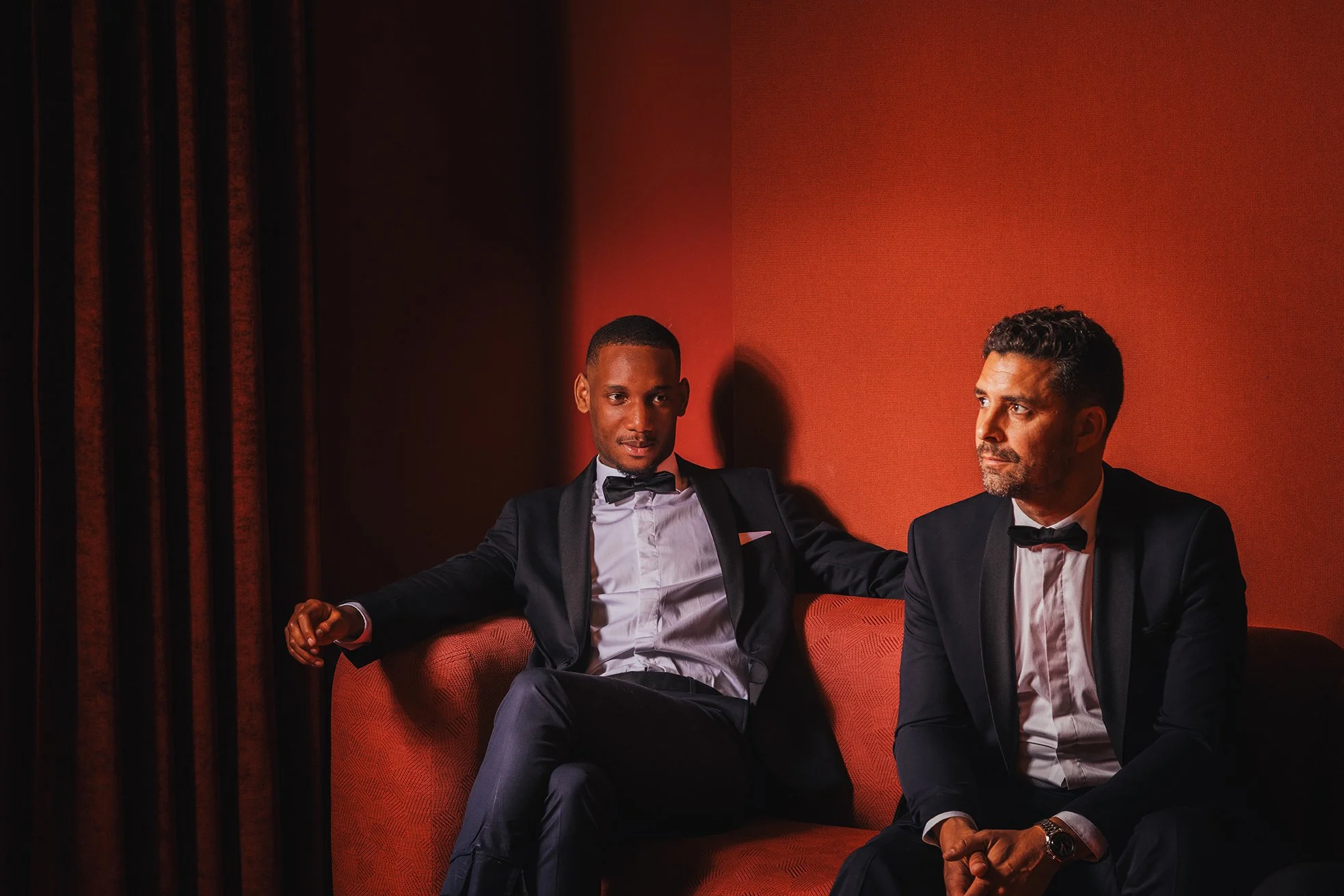 Deux hommes en costume noir et nœud papillon assis dans un fauteuil rouge contre un mur orange, avec une tenture en velours brun à gauche.
