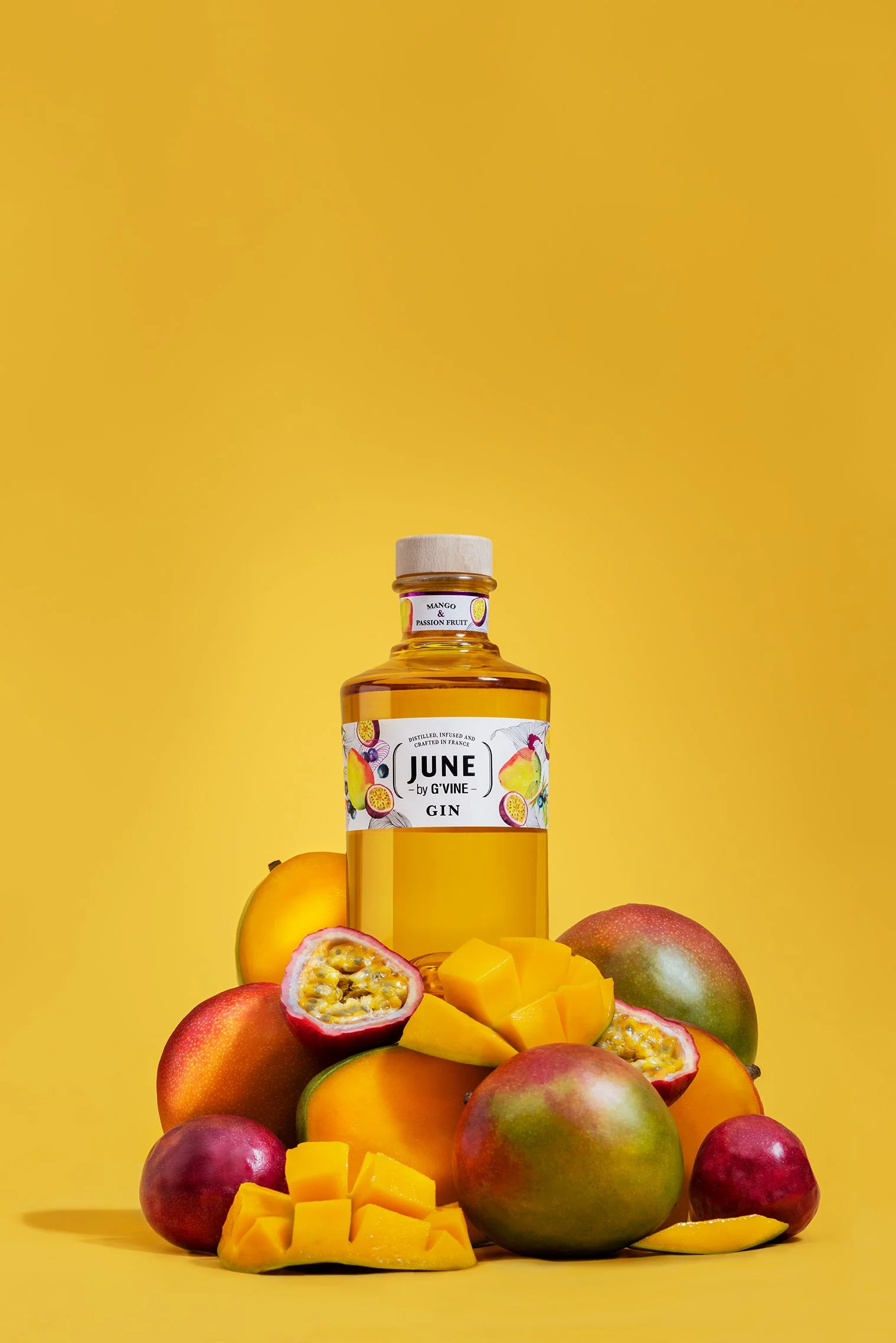 Une bouteille de gin IUNE à côté de mangues, papayes et nectarines, avec des cubes de mangue et des morceaux de fruits coupés, sur un fond jaune.