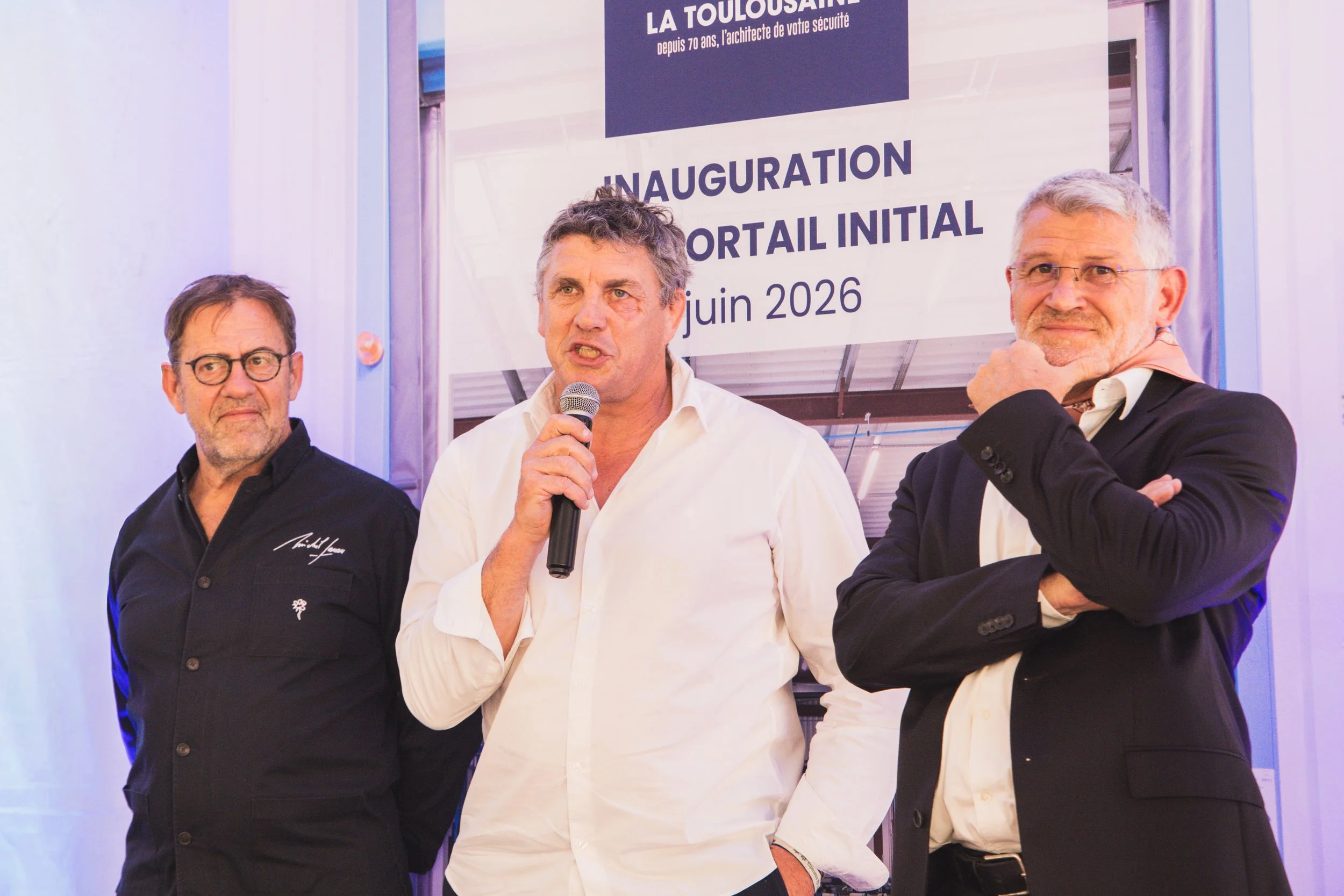 Trois hommes debout lors de l'inauguration d'une porte électrique à Toulouse, en juin 2026. Un homme au centre parle dans un microphone, tandis que les autres portent des costumes. Un panneau en arrière-plan affiche le texte ‘LA TOLOUSAINE’, ‘INaugur
