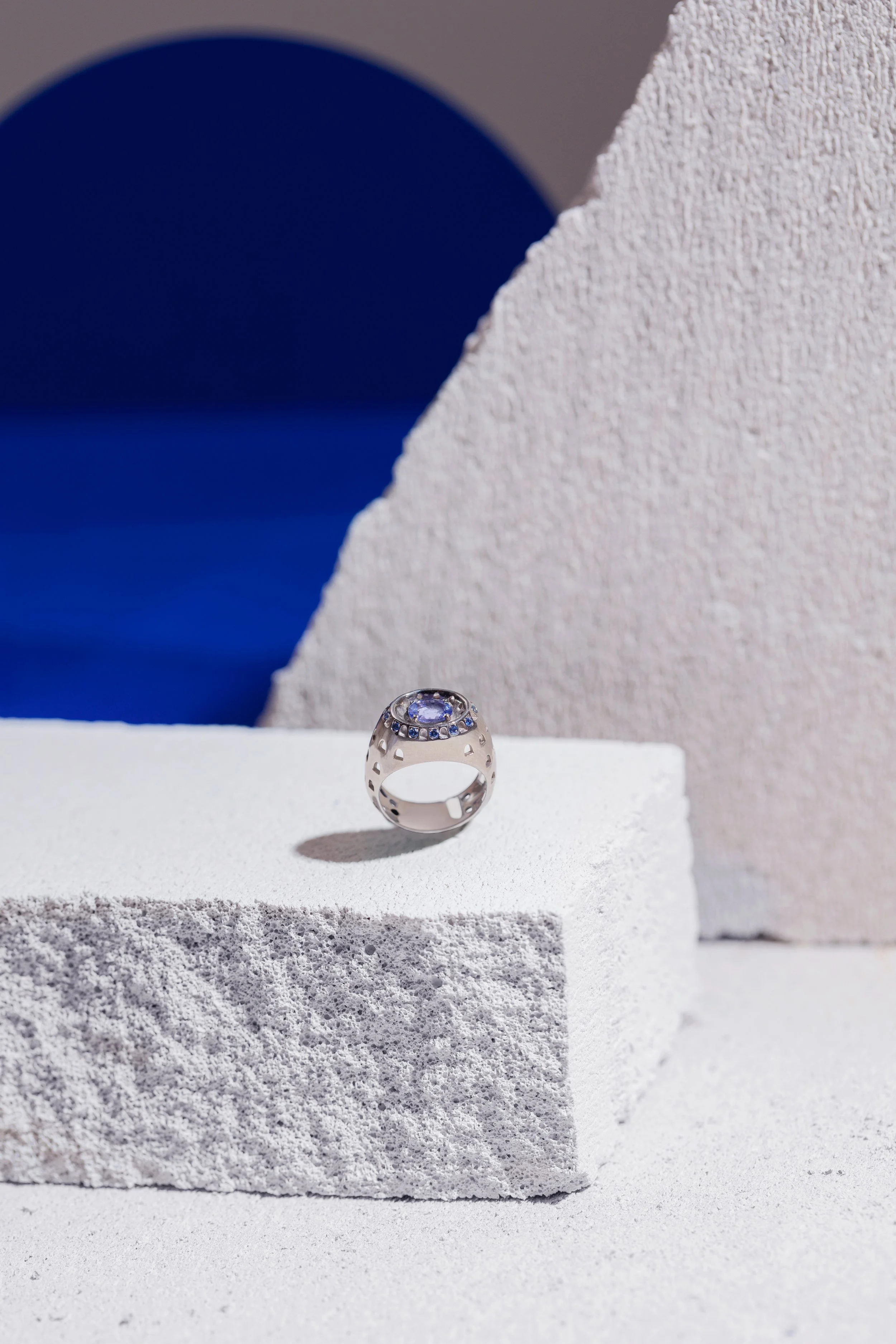 Bague en argent avec un grand saphir et des petites pierres incrustées, posée sur une surface blanche en terrazzo, avec un fond coloré de formes géométriques en blanc, beige, bleu et gris.