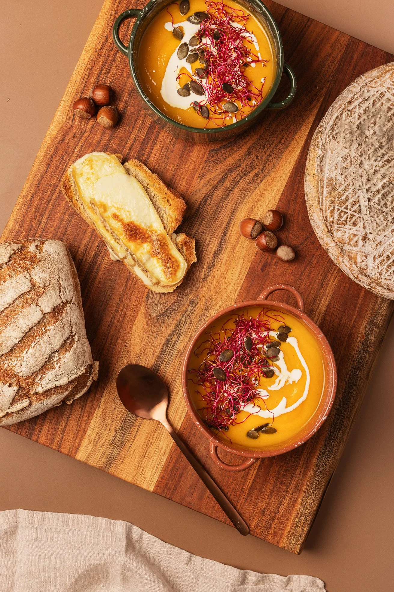 Soupe à la citrouille garnie de graines de courge, de pousses d'alfalfa et de crème, servie dans une casserole en terre cuite avec du pain grillé et des noisettes sur une planche en bois.