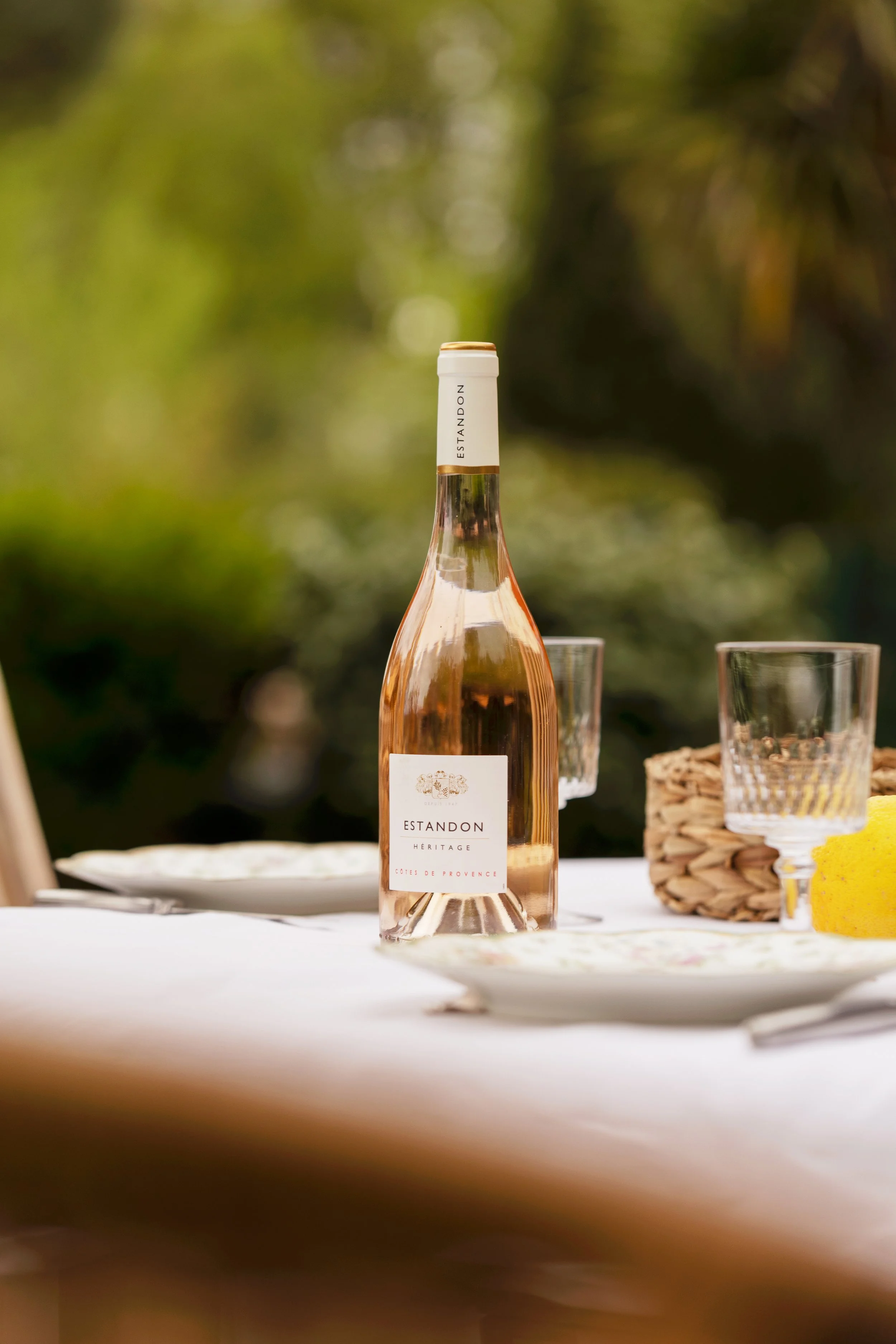 Bouteille de rosé rosé avec étiquette blanche portant l'inscription 'ESTANDON HERITAGE CÔTES DE PROVENCE', positionnée au centre, avec deux verres à côté, sur une table blanche, dans un environnement naturel avec un fond flou de verdure.