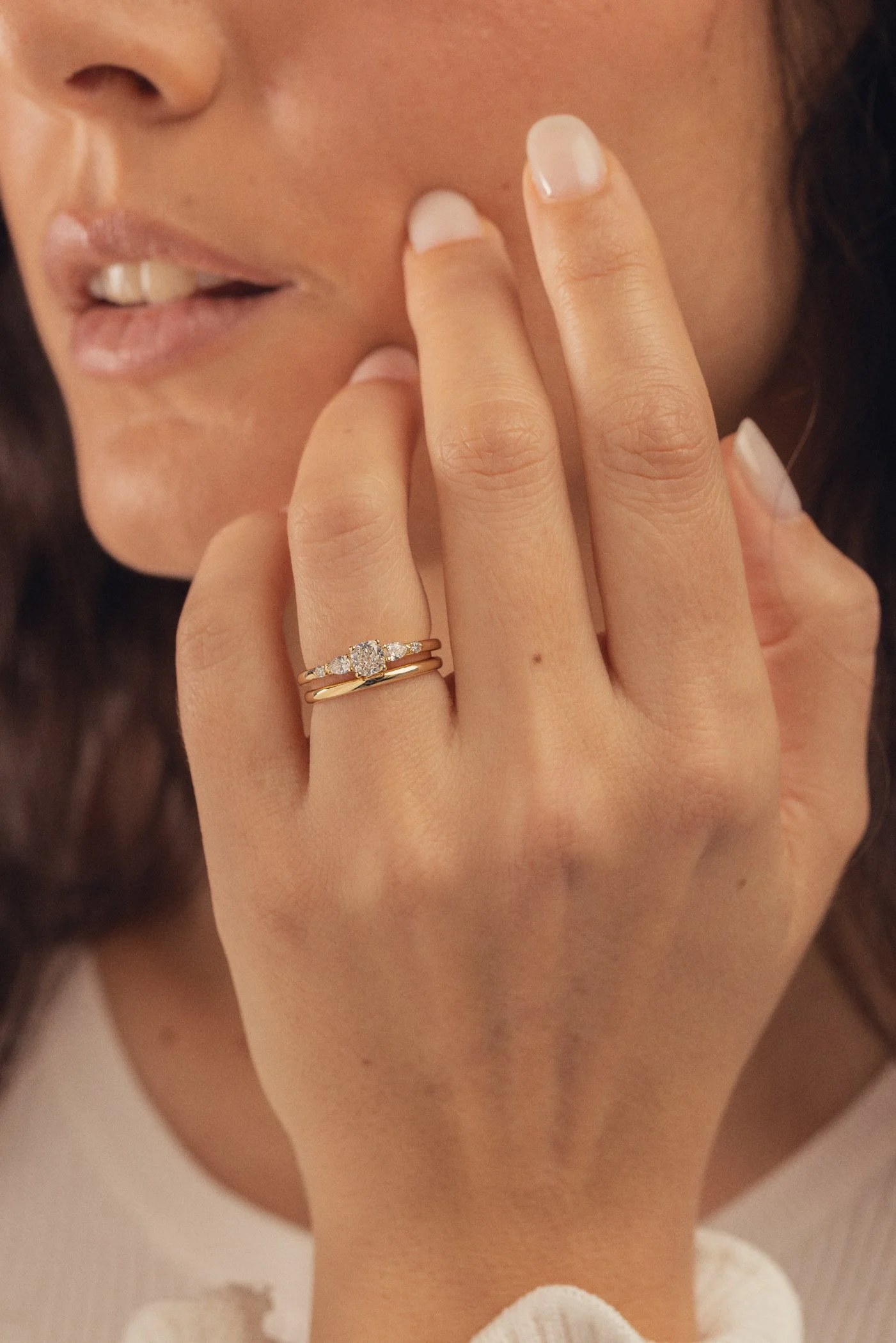 Femme portant une bague en or avec un diamant sur l'annulaire gauche, face à la caméra, mains près du visage, en train de toucher ses lèvres, cheveux bouclés, peau claire, sourcils bien dessinés.