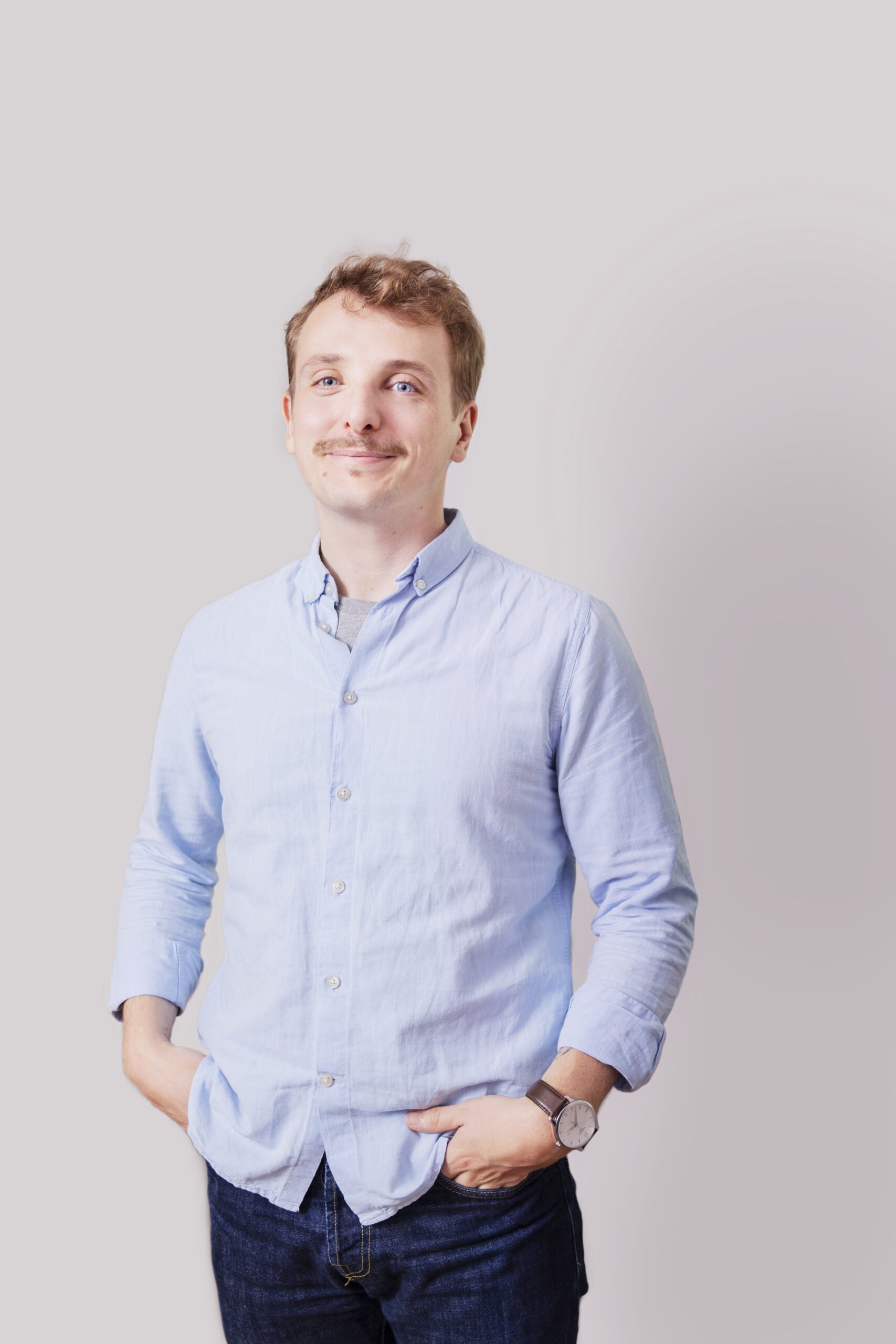 Jeune homme costumé en chemise bleu clair et jeans, posant contre un mur gris, souriant.
