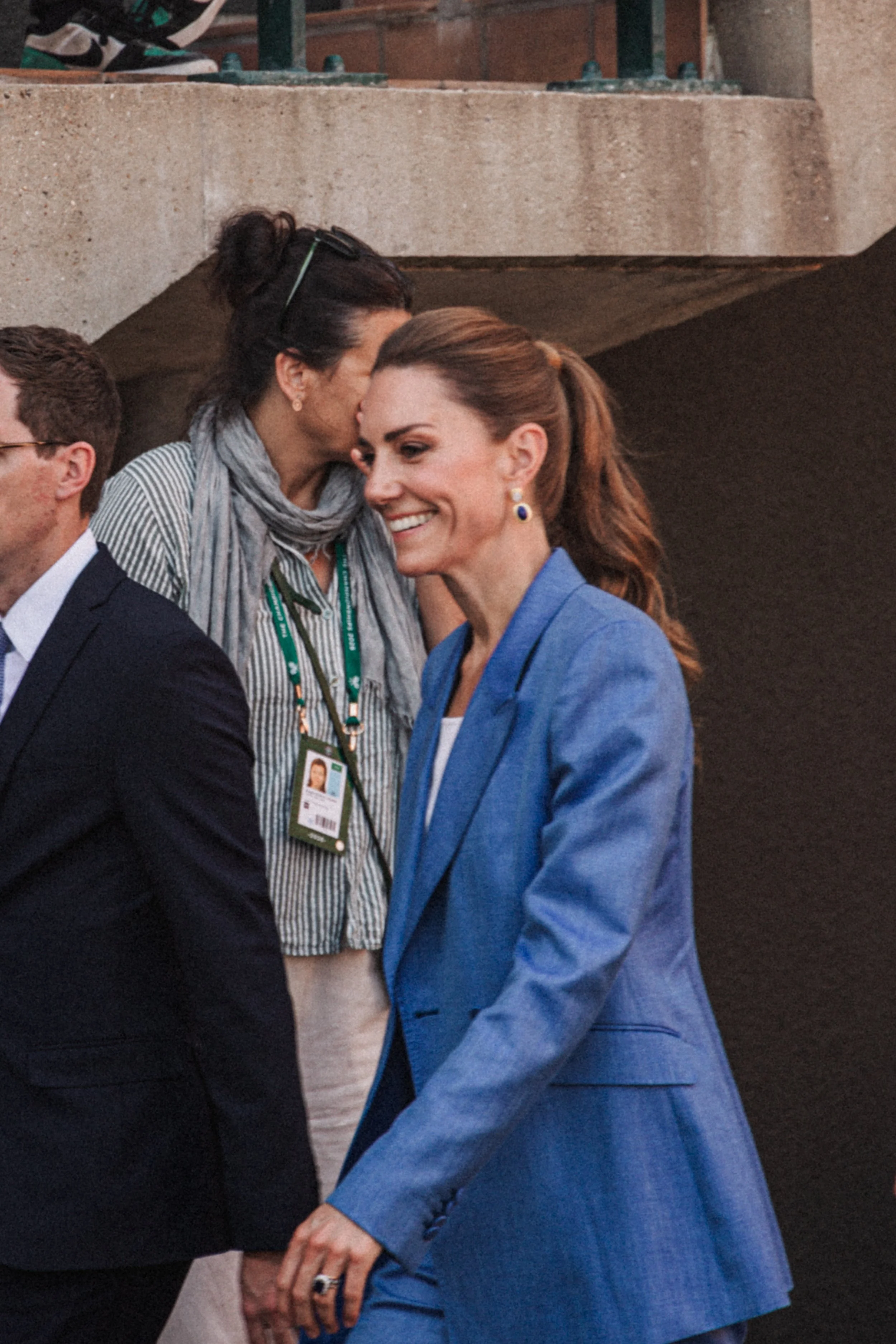 Deux femmes se tiennent la main et s'embrassent brièvement lors d'une rencontre publique. L'une porte un costume bleu, l'autre une veste sombre et possède un badge d'identification autour du cou.