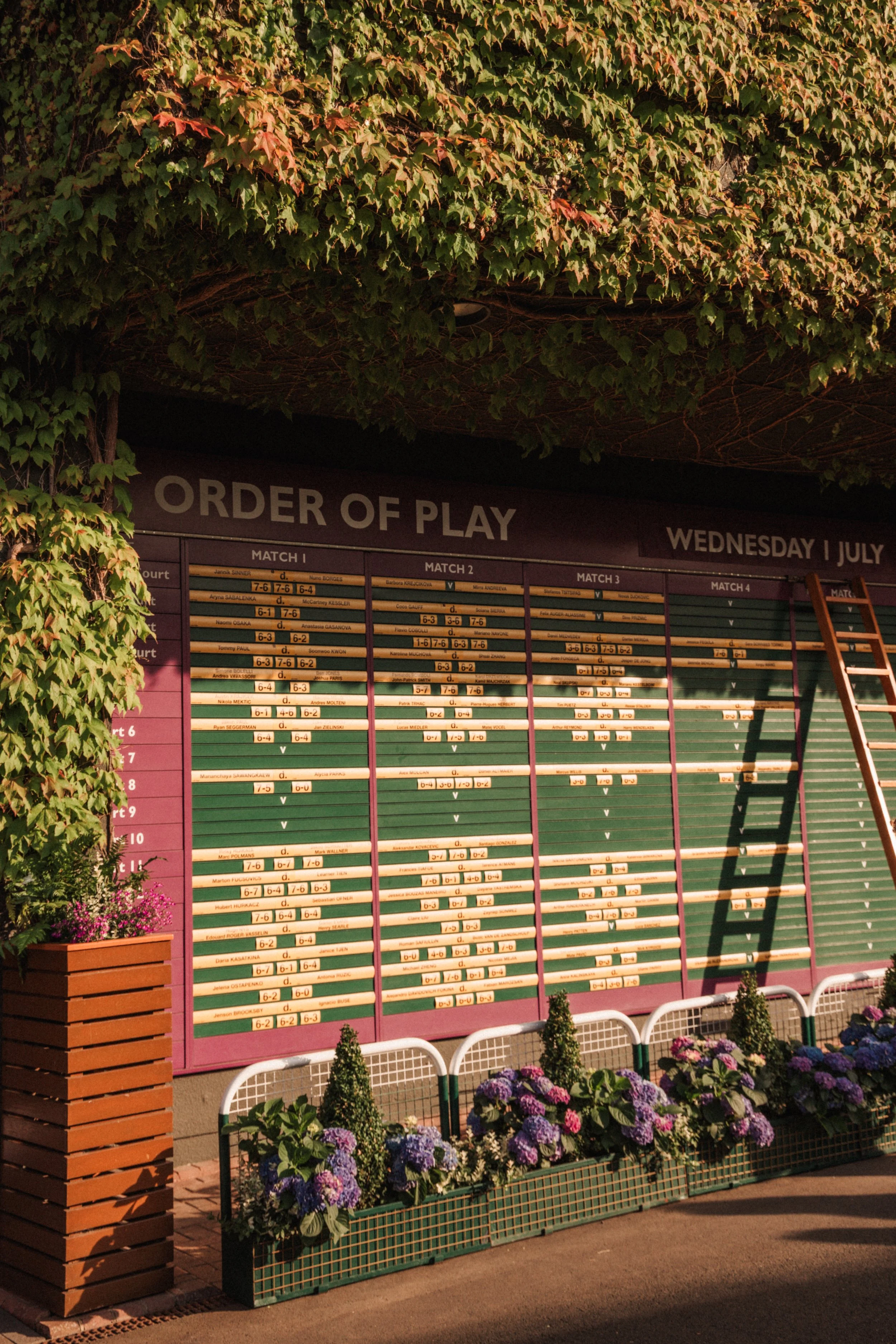 Tableau de résultats de matchs de tennis avec dates, noms des joueurs, scores et lettres de victoire, sous un panneau marqué 'ORDER OF PLAY' entre deux colonnes de fleurs dans un jardin.