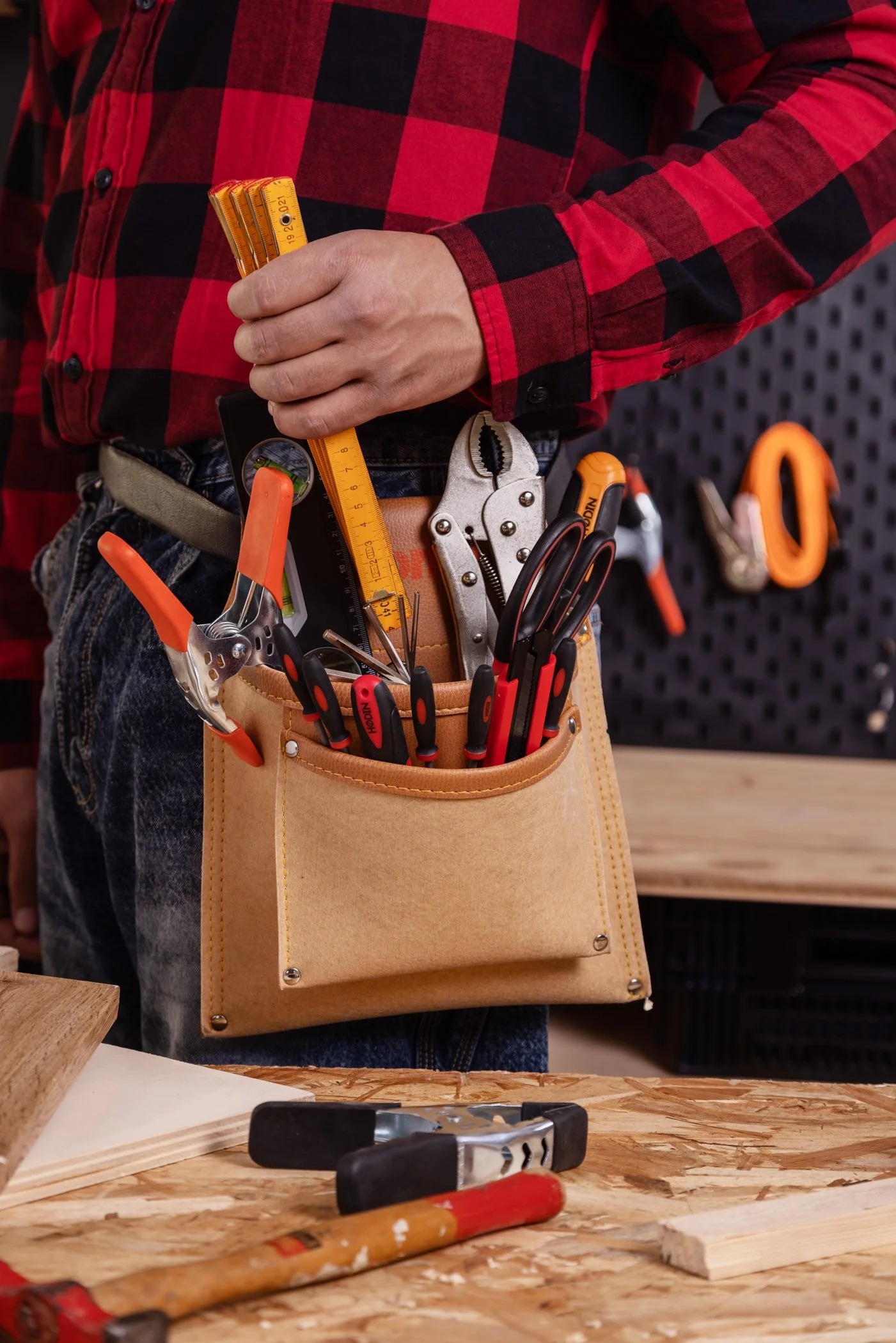 Un homme portant une chemise à carreaux rouges et noirs avec une ceinture en cuir, doté d'une ceinture à outils contenant des tournevis, une pince, un marteau et un mètre ruban, un atelier en arrière-plan avec un panneau perforé, des outils suspendus.