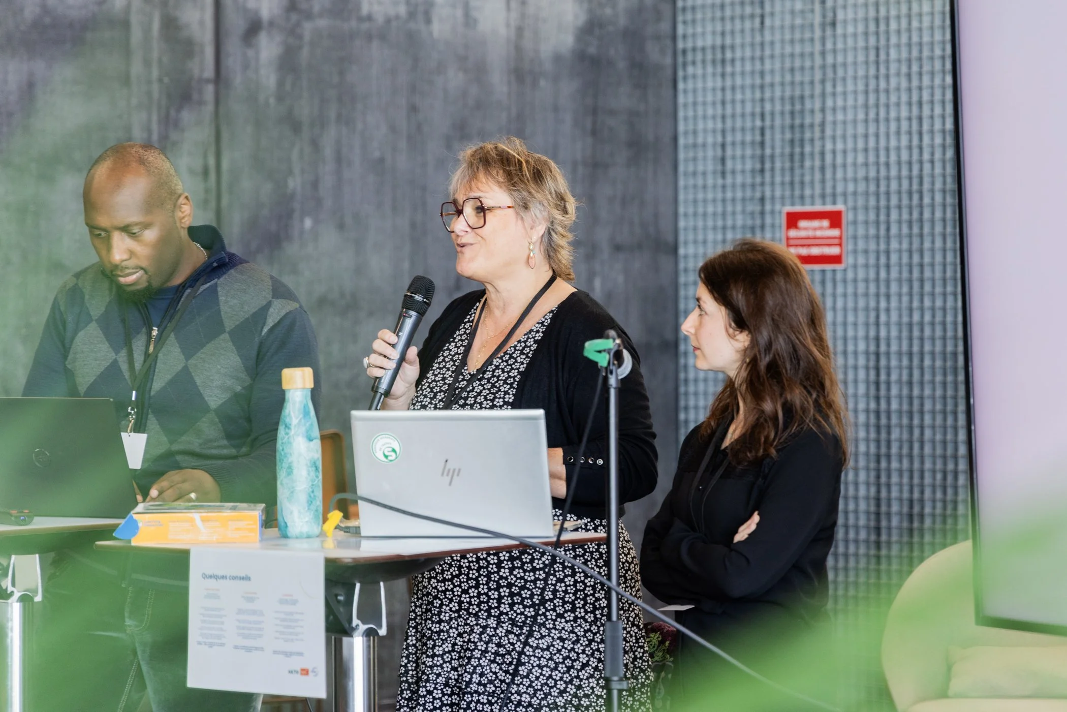 Une femme âgée avec des lunettes parle dans un micro lors d'une réunion ou conférence, entourée de deux autres personnes, dans un environnement intérieur moderne.
