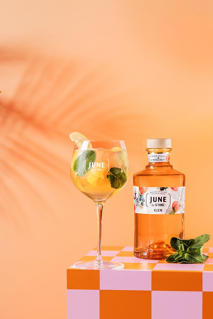 Une bouteille de gin rose sur une table à carreaux oranges et blanches, à côté d'un verre de cocktail avec des feuilles de menthe et des tranches de citron, sur fond peach.
