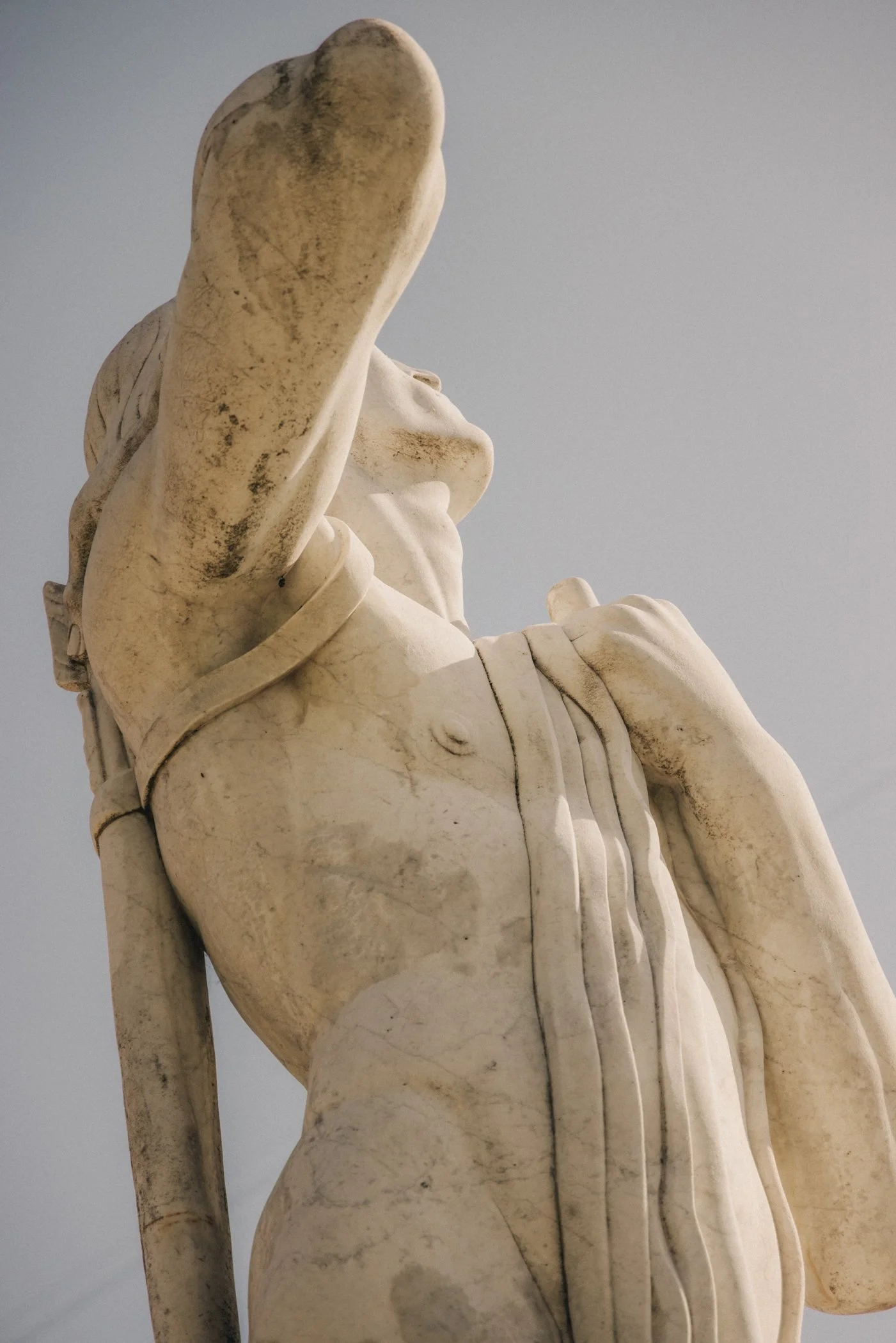 Statue en marbre blanc représentant une femme nue, vue de profil, avec la main levée pour couvrir la face, contre un ciel gris.
