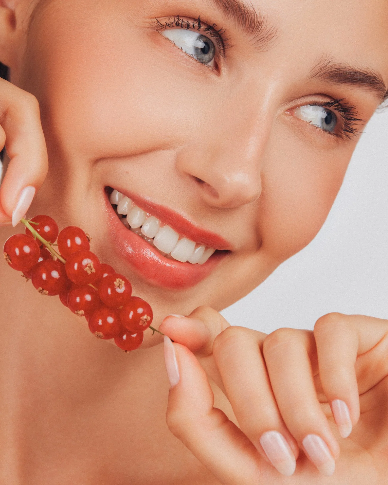 Un gros plan d'une femme souriante avec des yeux bleus, tenant une branche de groseilles rouges près de sa joue, avec des ongles soignés et un maquillage léger.