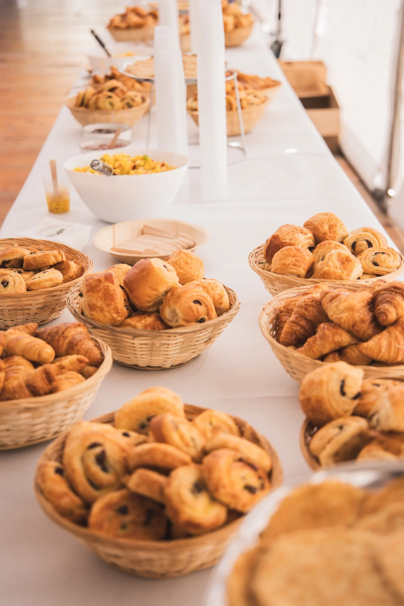 Table avec des paniers de viennoiseries telles que des croissants et des pains au chocolat, accompagnés de bols avec des confitures ou des accompagnements. En arrière-plan, des assiettes avec des tartes ou des quiches.
