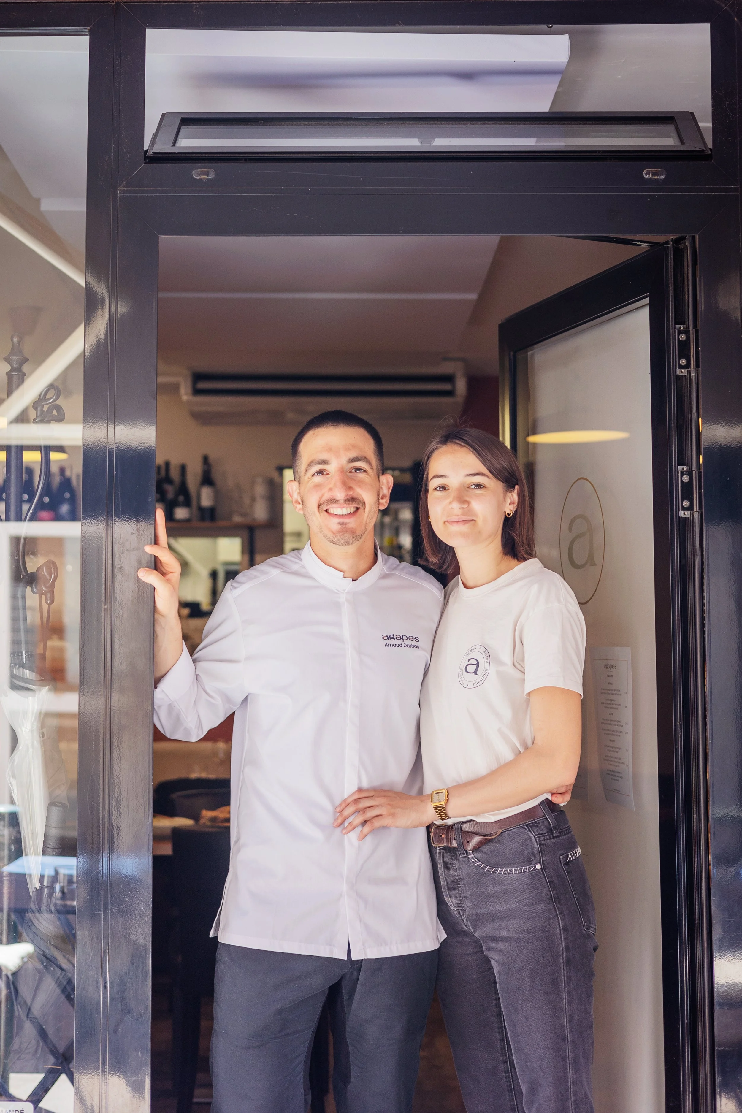 Deux personnes, un homme en uniforme de cuisine et une femme en vêtements décontractés, posent côte à côte dans une porte d'entrée d'un restaurant ou café, souriant à la caméra.
