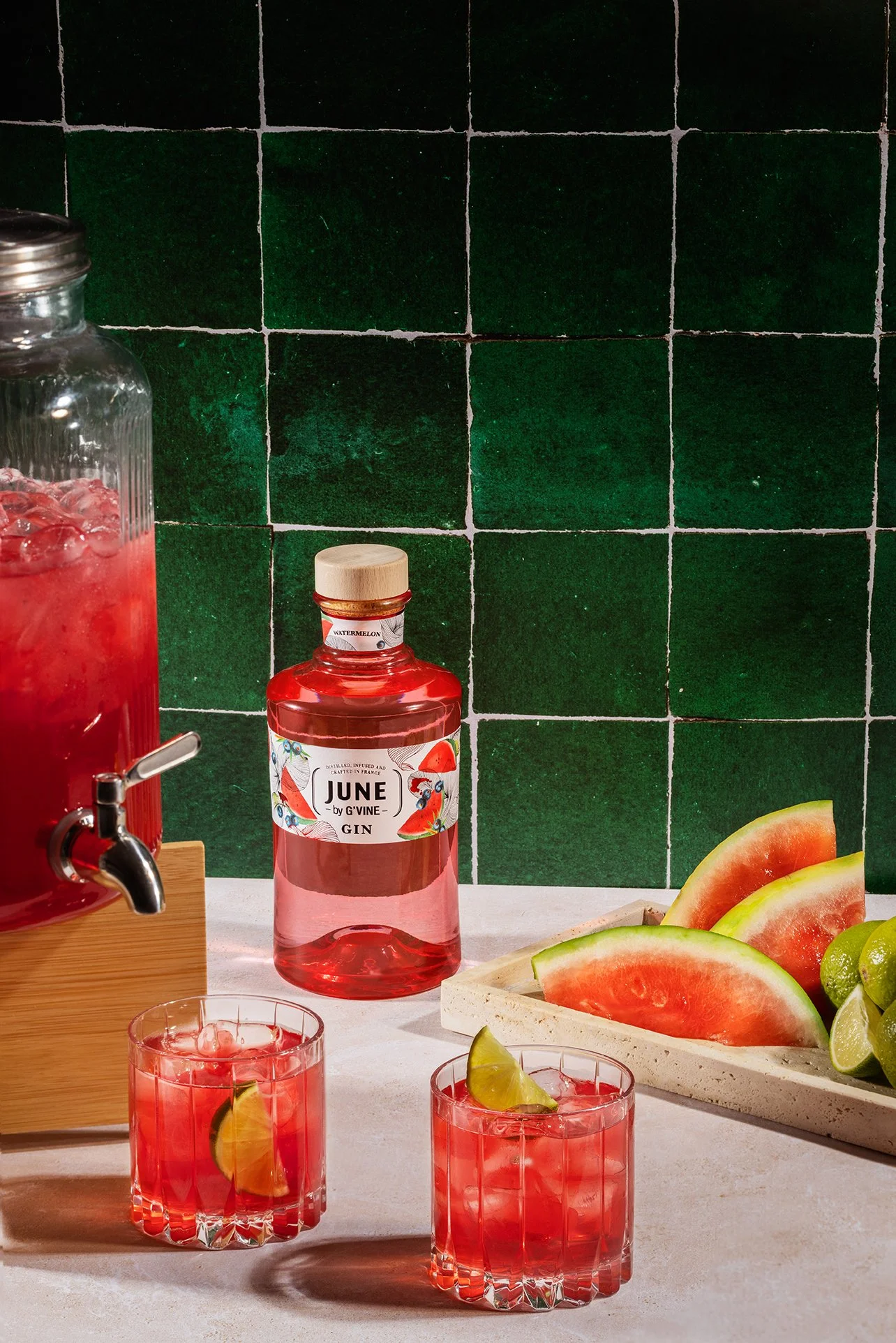 Verre de boisson rose avec une tranche de citron, tranche de pastèque, et tranche de lime. Plateau avec des tranches de pastèque, citron et lime. Bouteille de gin aromatisé à la pastèque, avec un fond de carreaux verts foncés.