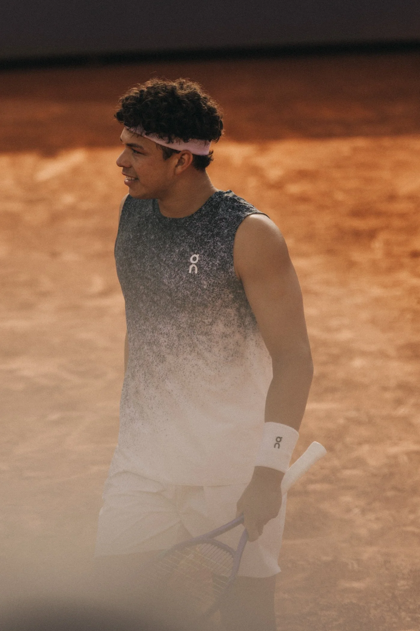 Un jeune homme avec des cheveux bouclés portant un bandeau rose, une tenue de sport grise et blanc, tenant une raquette de tennis, sur un court de terre battue