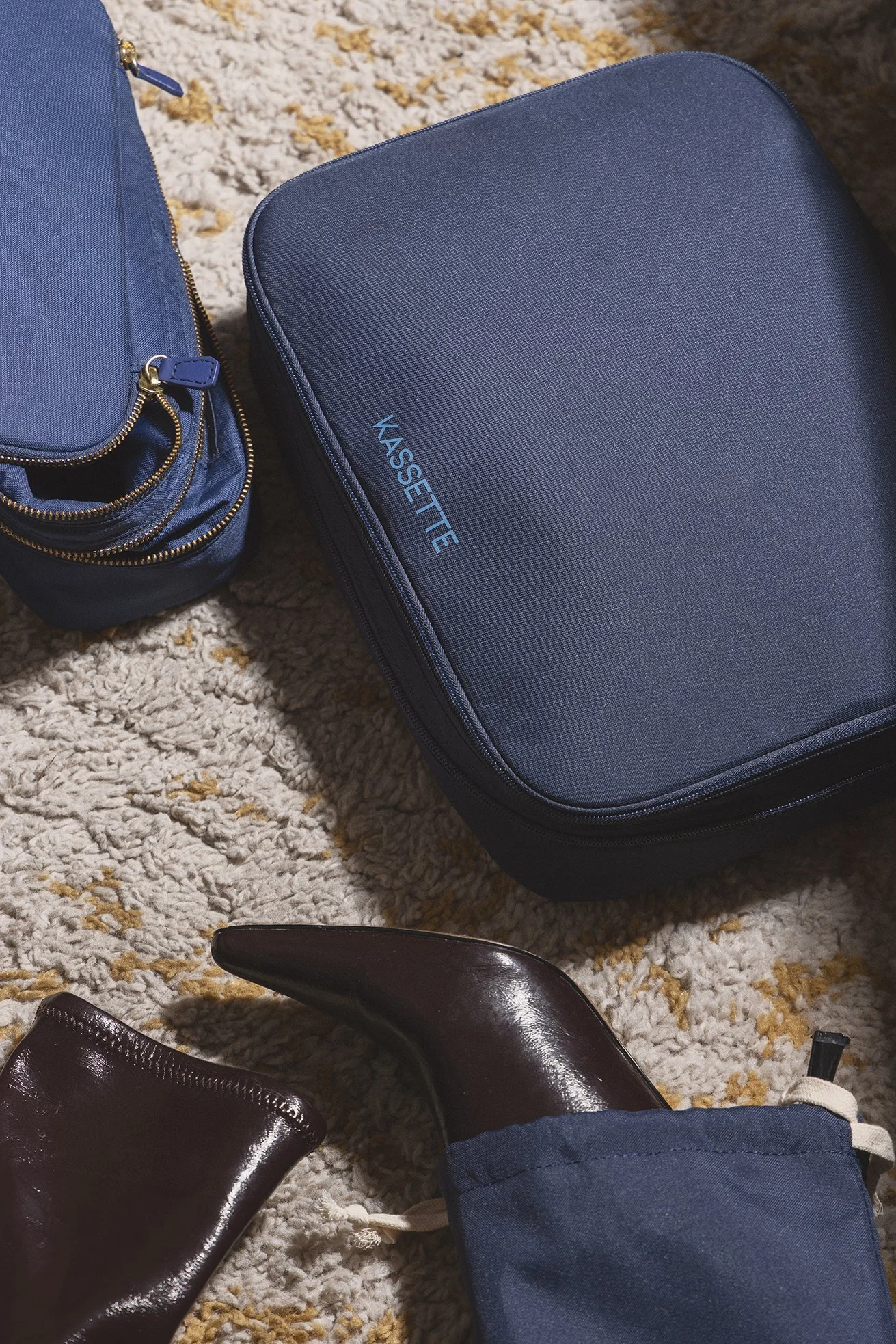 Une photo de plusieurs sacs et chaussures sur un tapis moelleux, notamment un sac à dos bleu, une valise bleue, une paire de chaussures en cuir marron, et une chaussure beige à la marche. On voit aussi une chemise ou pantalon en jean bleu.