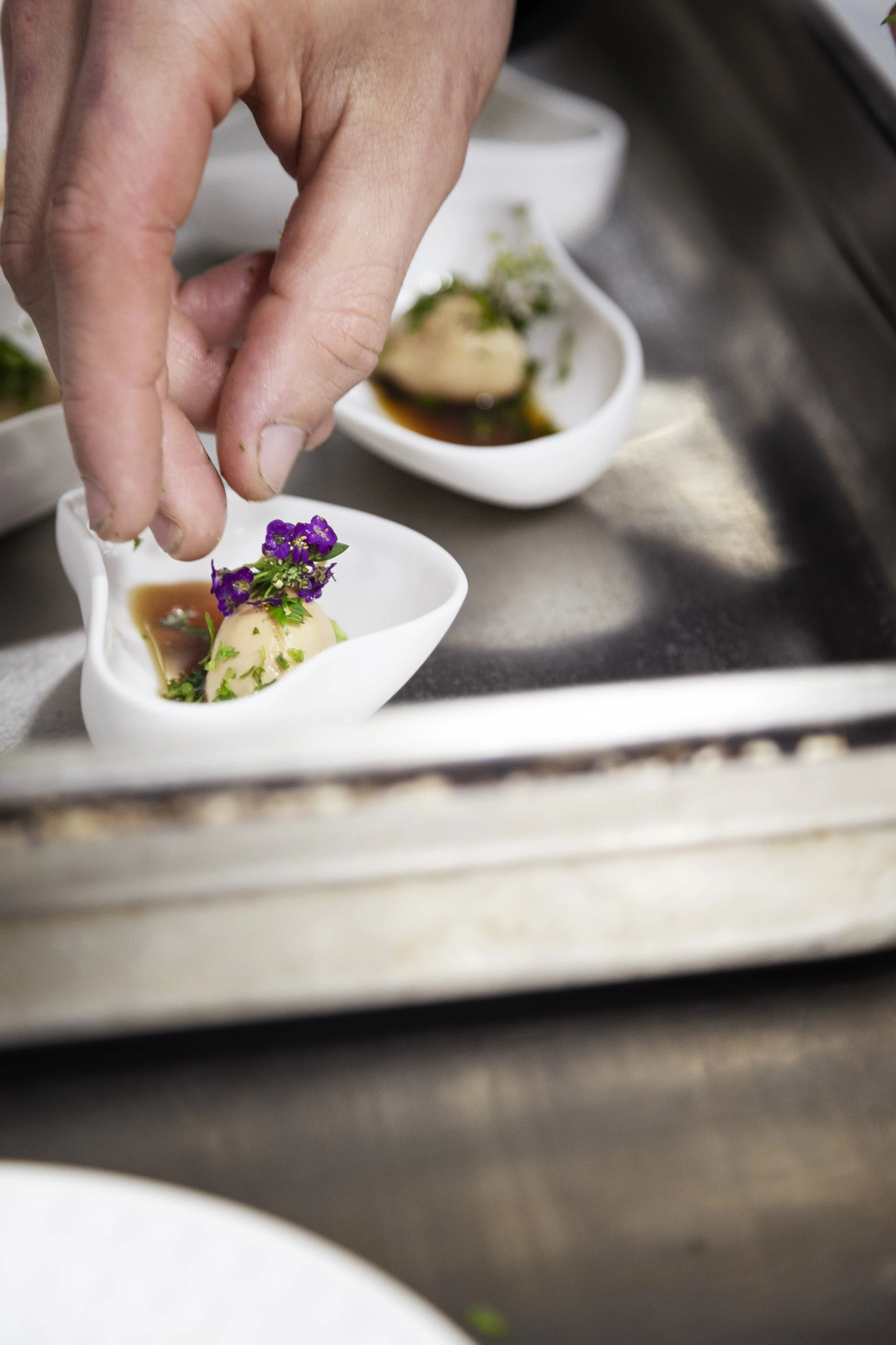 Une main saupoudre des fleurs violettes sur un plat de nourriture dans une petite assiette blanche.