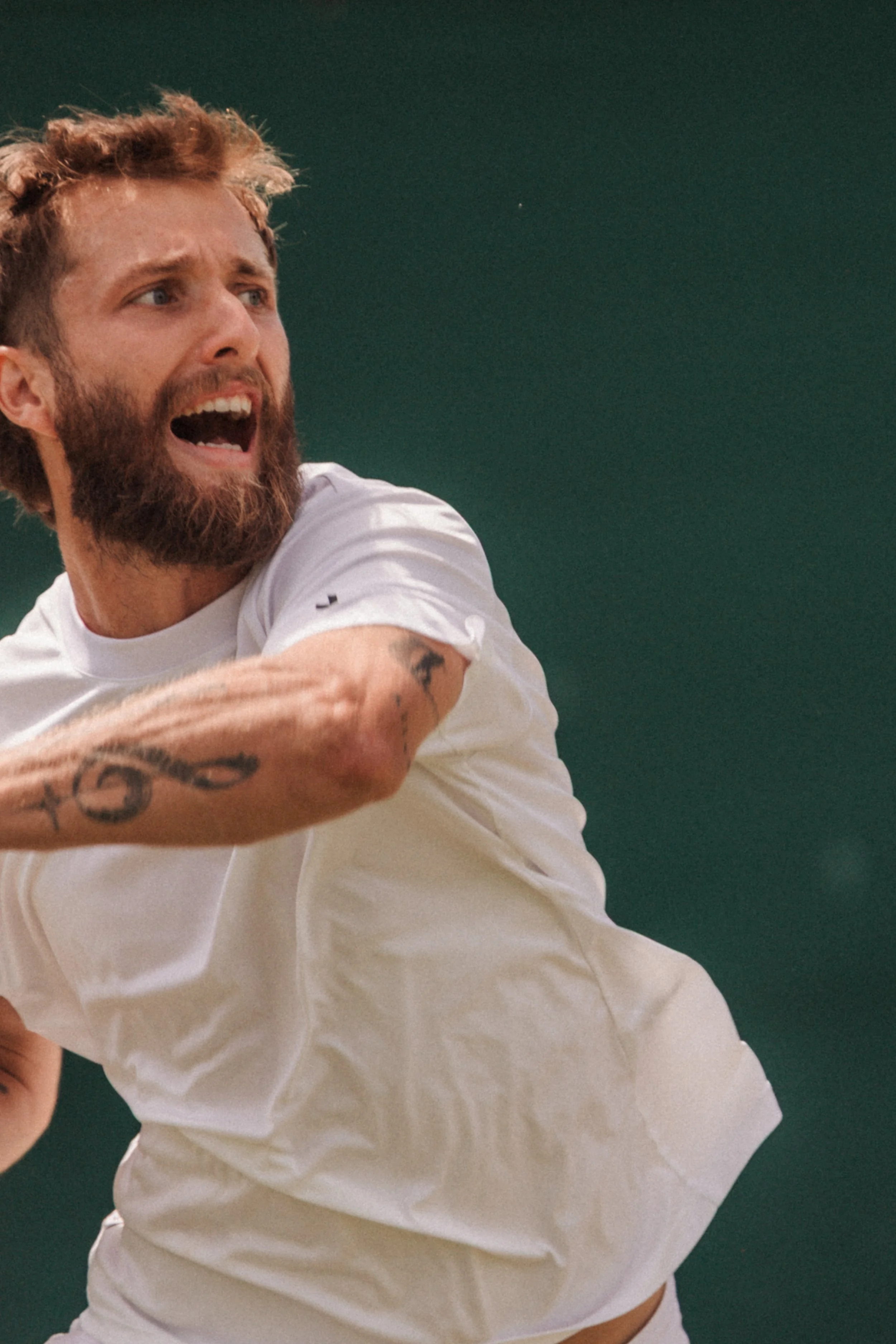 Homme avec une barbe, en train de faire un geste de combat ou de performance sportive, portant un t-shirt blanc, sur un fond vert.