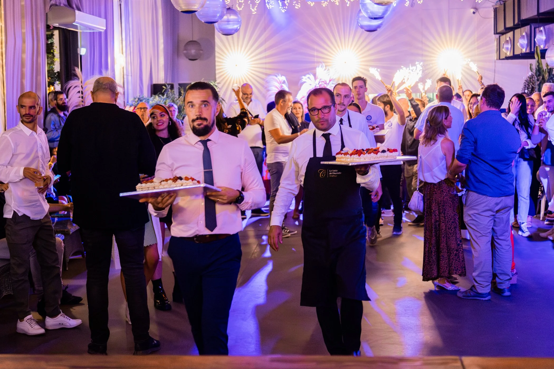 Une foule de personnes lors d'une fête avec des serveurs portant des plateaux de desserts et un décor coloré avec des lumières et des confettis.