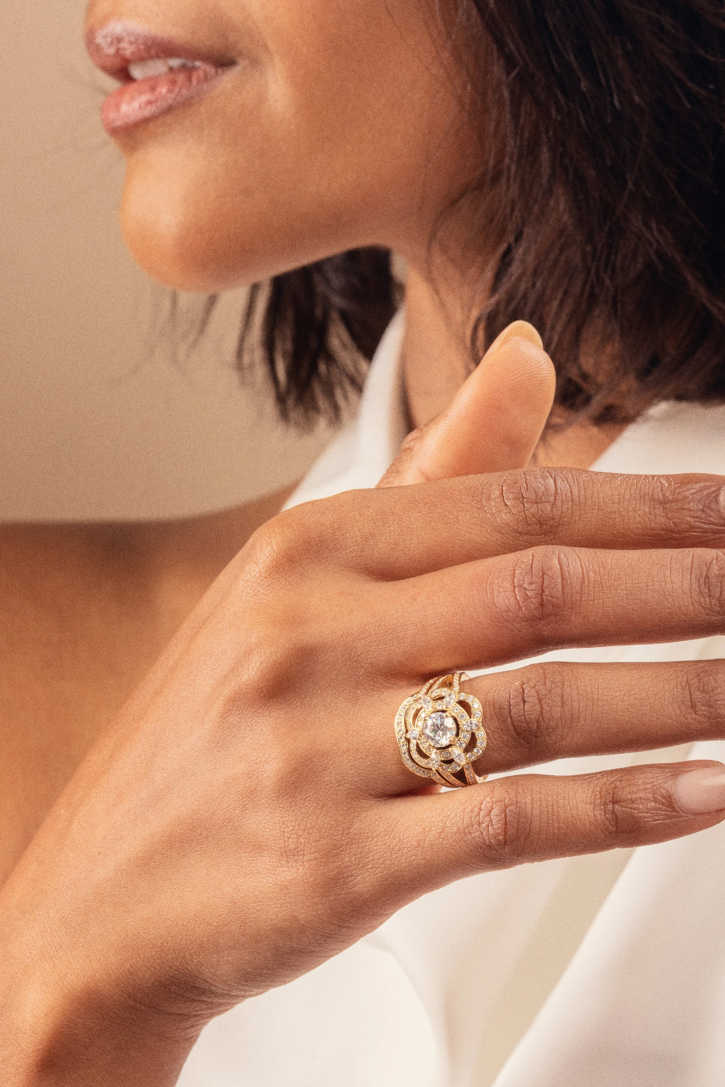 Une main féminine montre un anneau en or orné de diamants sur son doigt. La femme a une peau de couleur moyenne et porte une bague élégante. Elle a également des cheveux bruns foncés et porte un maquillage minimaliste. La photographie est une close-up détaillée de ses doigts et de son visage partiellement visible en arrière-plan.