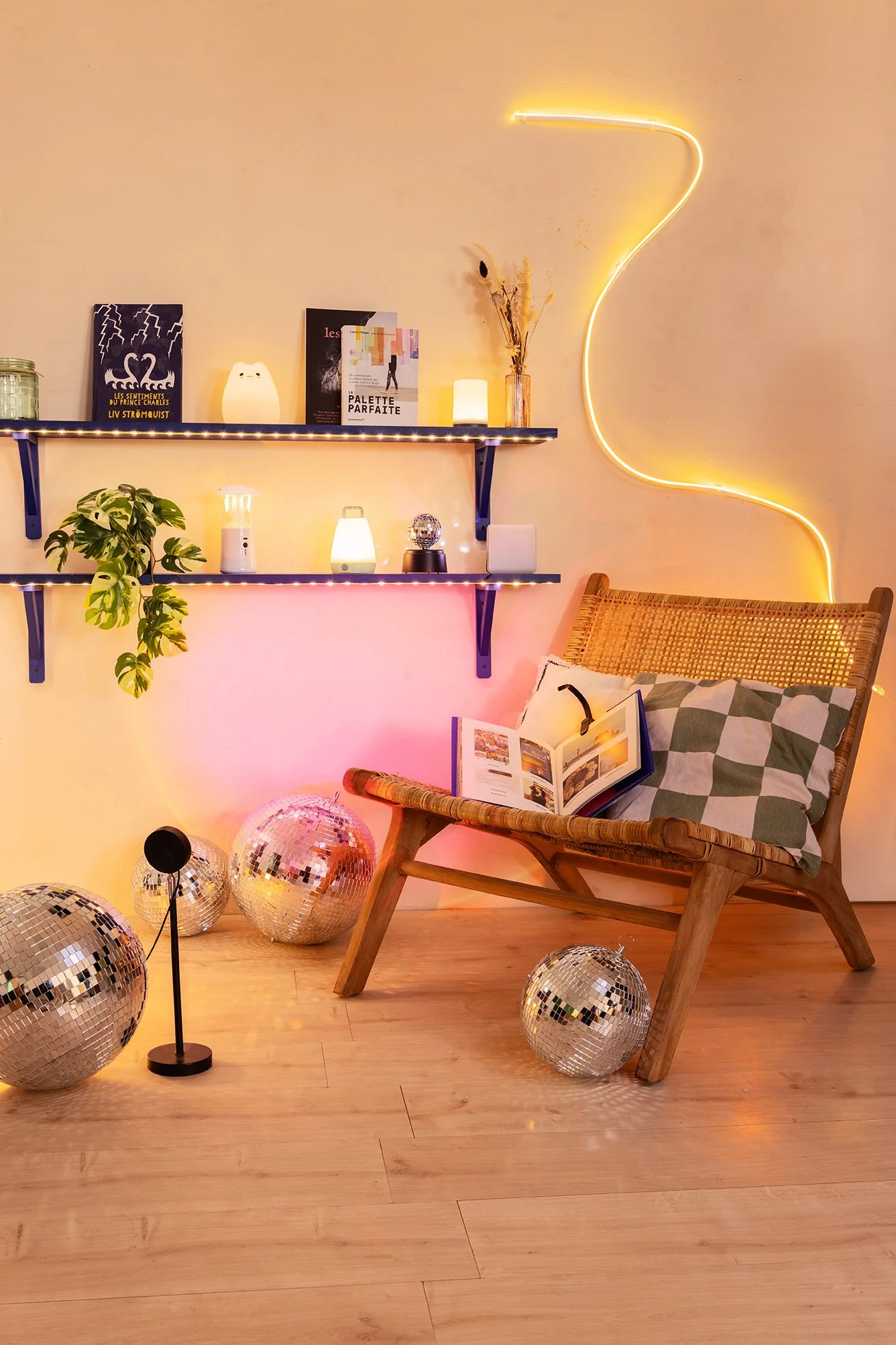 Un coin de chambre décoré avec des lampes LED, des livres, une plante, un fauteuil en rotin avec coussin à motif, des boules disco et une guirlande lumineuse en forme de courbe sur le mur.