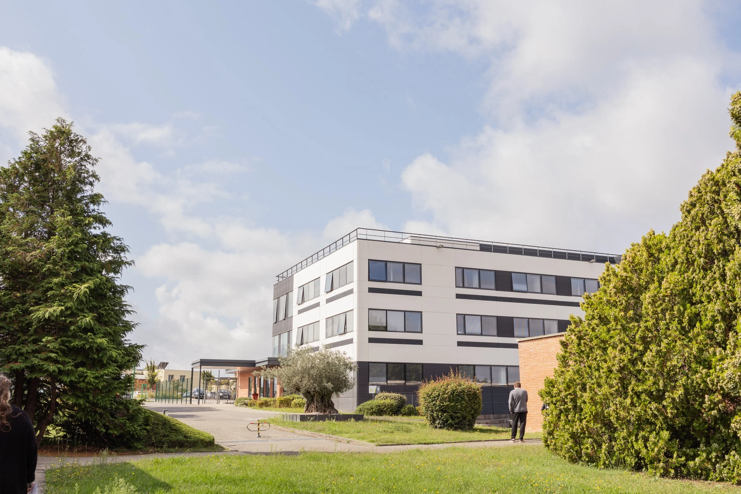 Un bâtiment moderne avec plusieurs fenêtres, entouré d'arbres et d'une zone herbeuse, sous un ciel partiellement nuageux.