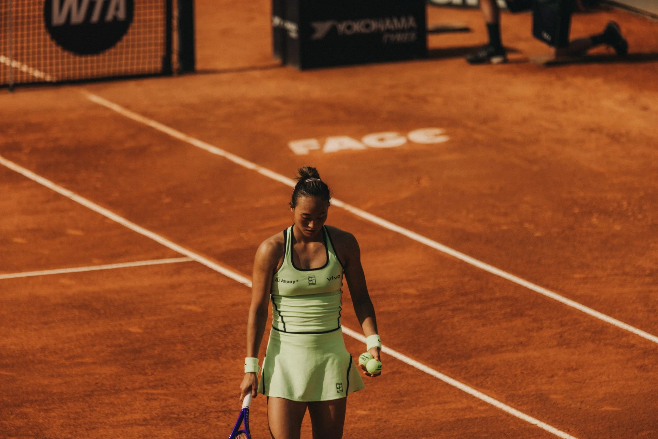 Une athlète féminine portant une tenue de sport verte, tenant une balle de tennis, se trouvant sur un court en terre battue, avec des équipements sportifs et des affiches en arrière-plan.