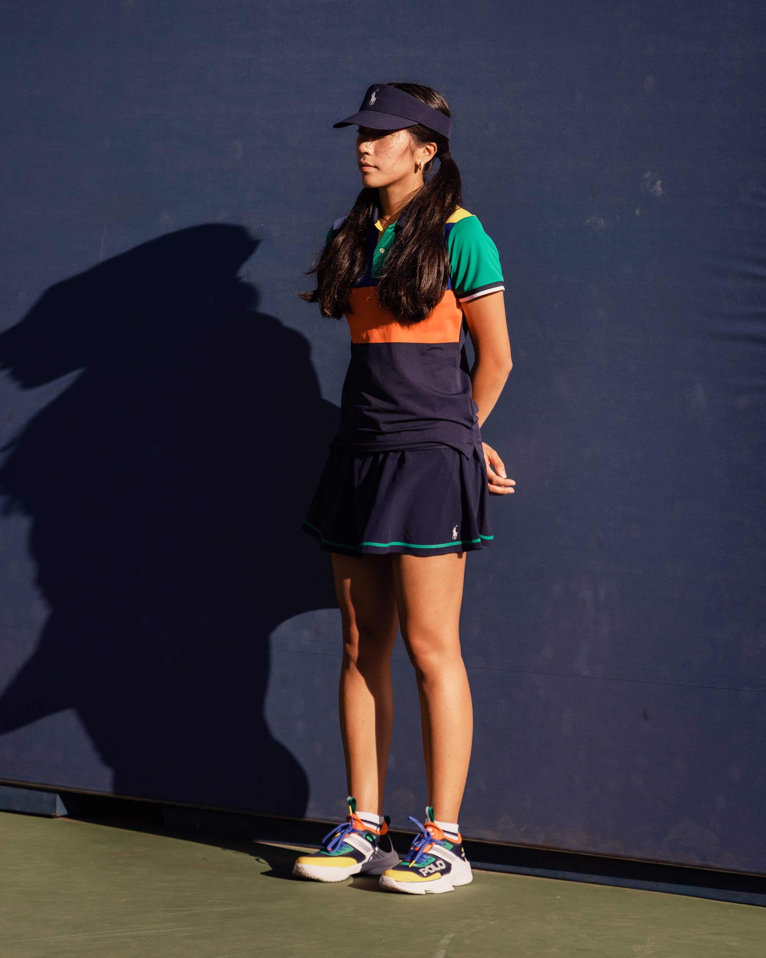 Une femme en tenue de sport debout sur un terrain de tennis, portant une casquette noire, un polo coloré, une jupe noire et des baskets. Son visage est partiellement ombragé par la casquette et elle regarde vers la droite, avec un fond sombre.