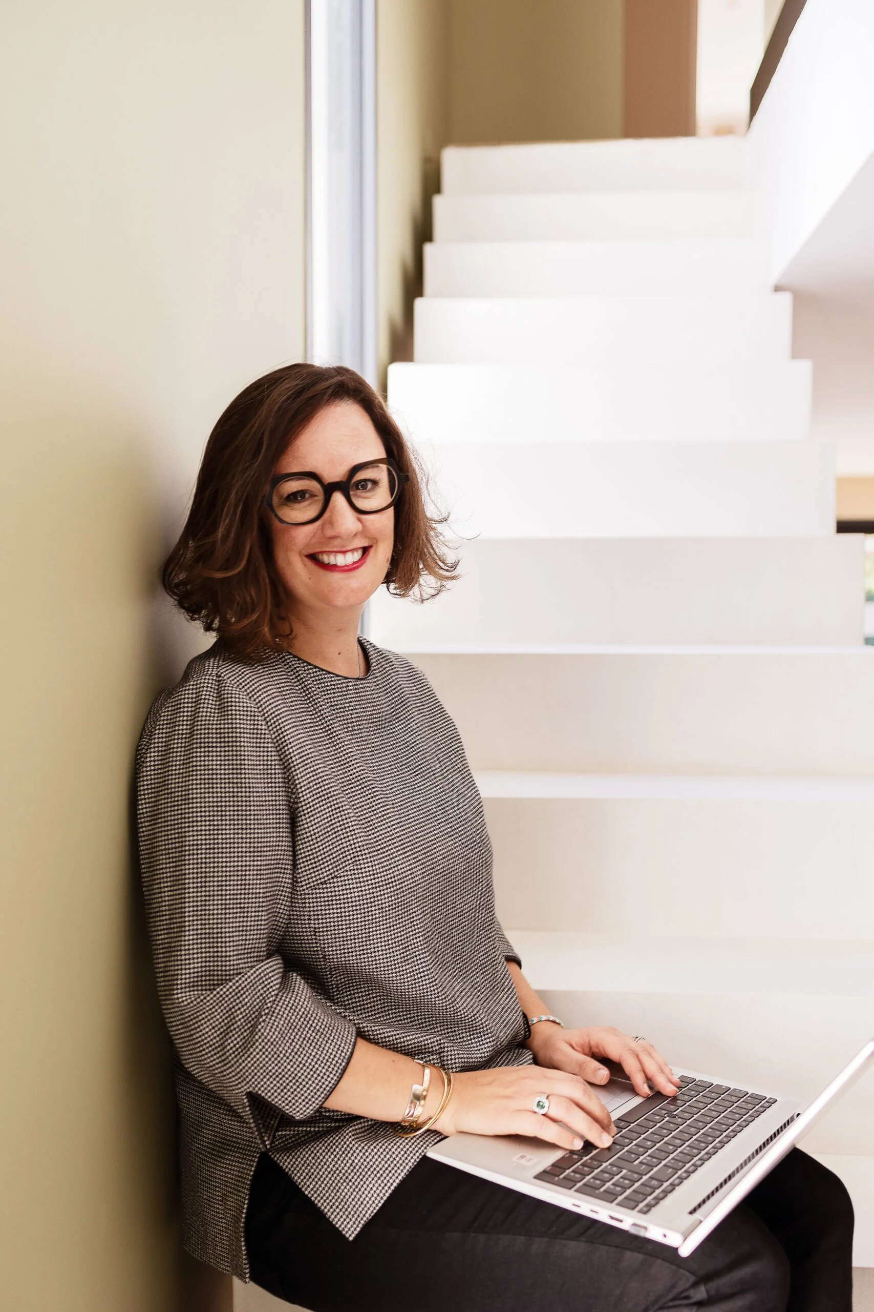 Une femme souriante portant des lunettes noires, avec des cheveux bruns, assise contre un mur vert clair, utilisant un ordinateur portable. En arrière-plan, il y a un escalier blanc en forme de zigzag.