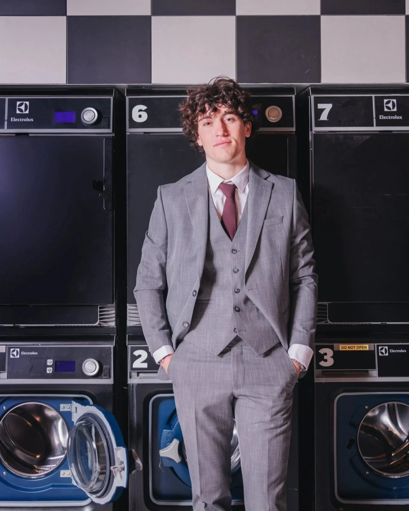 Un jeune homme en costume gris est debout dans une laverie automatique, parmi des machines à laver. Il a les mains dans les poches et regarde l'appareil photo.