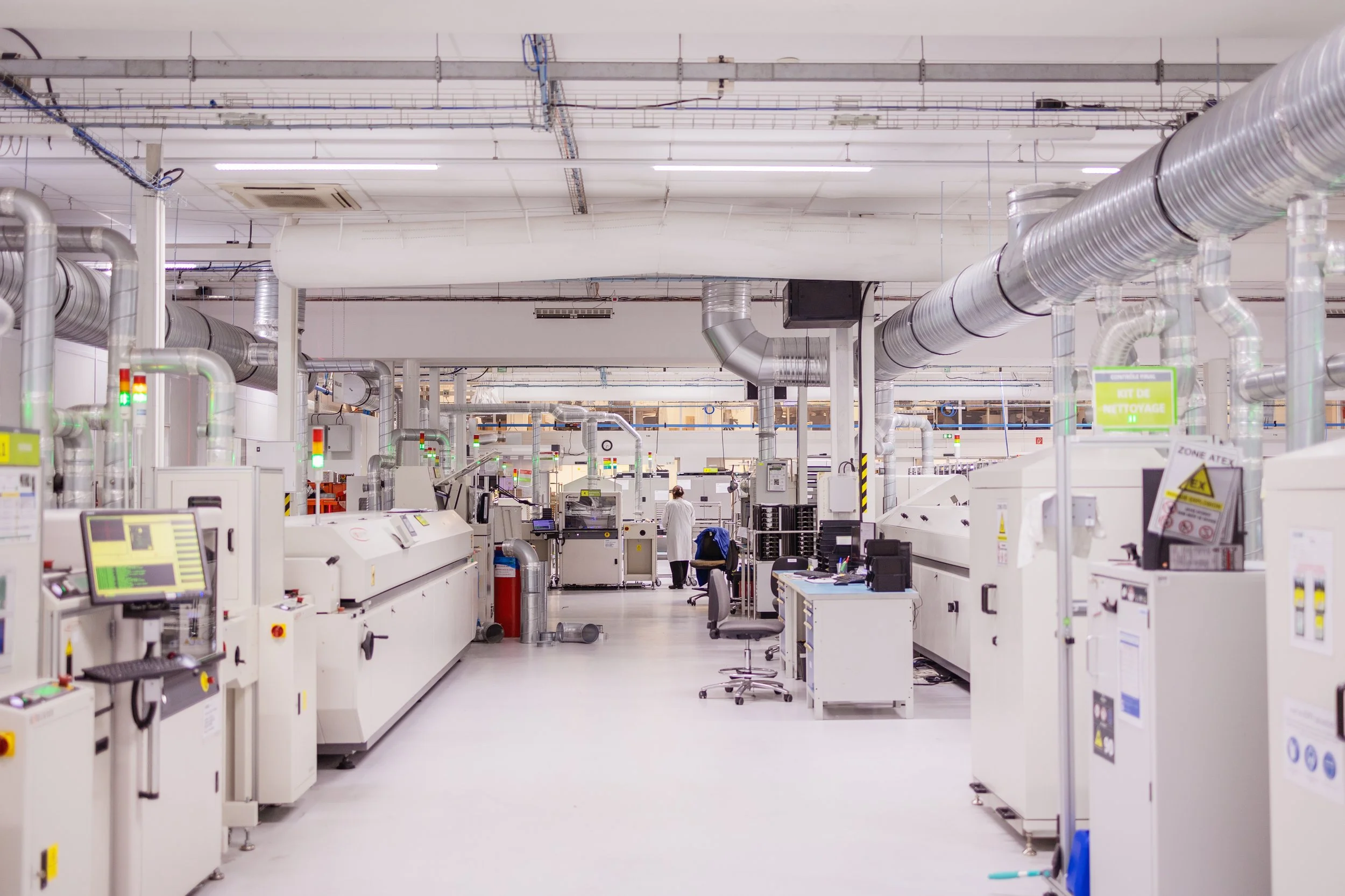 Vue d'une usine ou d'une salle de fabrication avec de nombreuses machines et conduits en métal, plusieurs employés en blouse travaillant à leur poste.